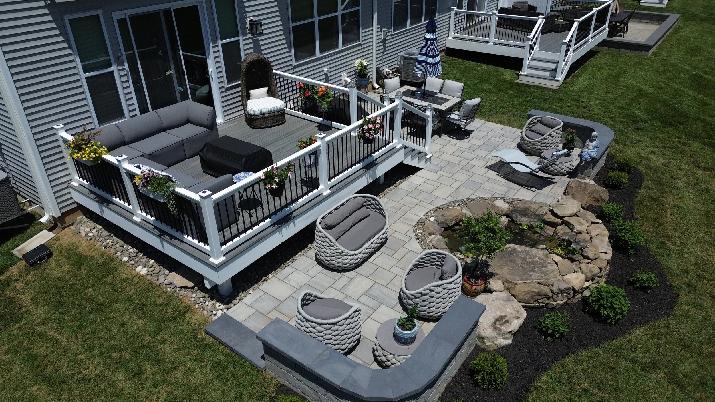 A backyard patio with outdoor furniture, including a cushioned sectional sofa, round lounge chairs, and a dining table with chairs, surrounded by a garden with rocks and green plants, and a deck with stairs attached to a house with horizontal siding.