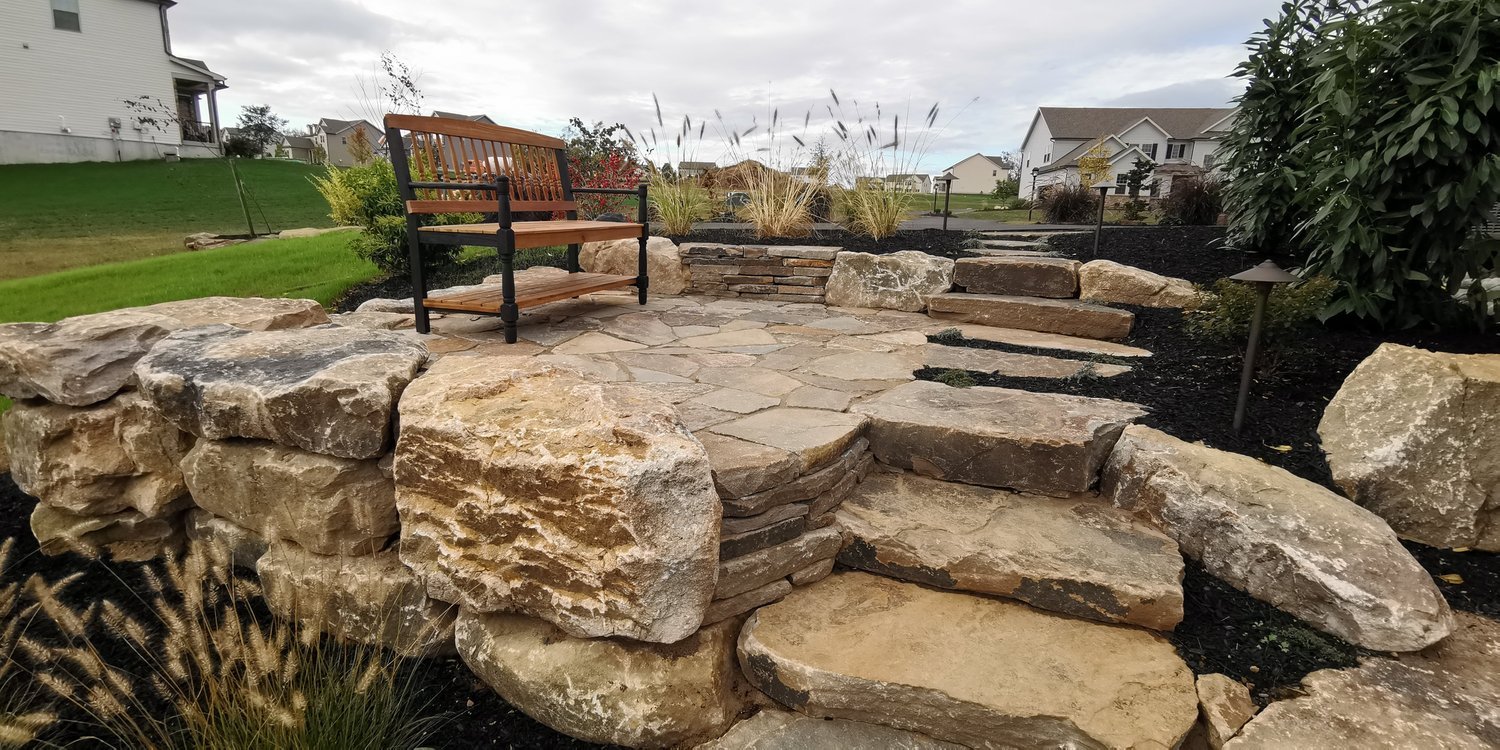 A landscaped backyard with a stone patio, stone steps, large rocks, a wooden bench, garden plants, black mulch, and surrounding suburban houses.
