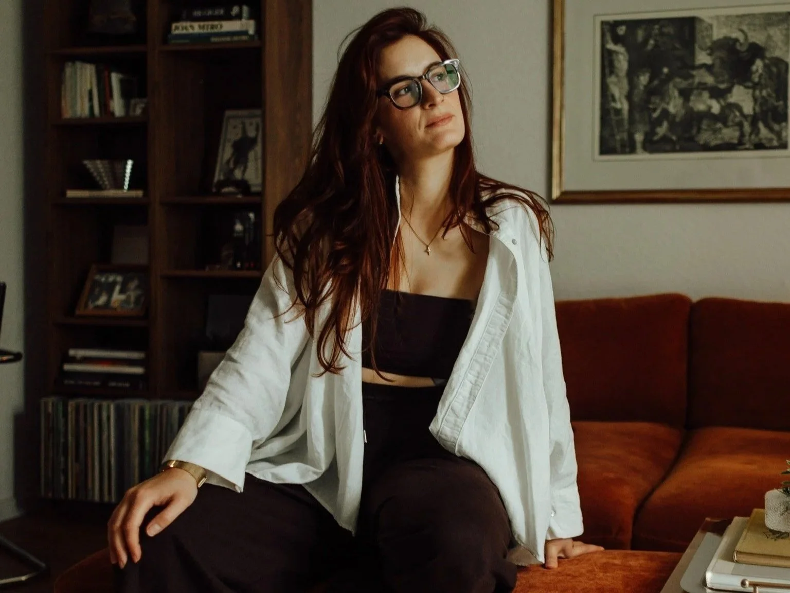 Julia Heeke, Interior Designerin, sitzt entspannt auf einem Hocker vor einem rostfarbenen MidCentury Sofa – im Hintergrund ein dunkles Bücherregal mit Kunstbüchern und eine gerahmte Grafik an der Wand.