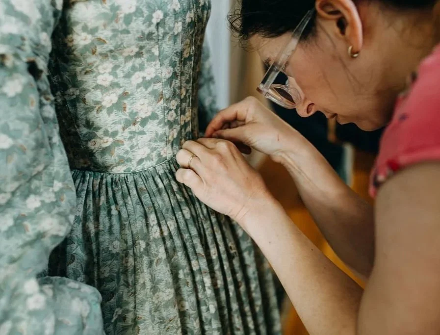 Image shows waist, upper skirt, and sleeve of green floral-printed dress from 1850s along with arms and head of fashion historian Whitney Robertson who is adjusting the waist.