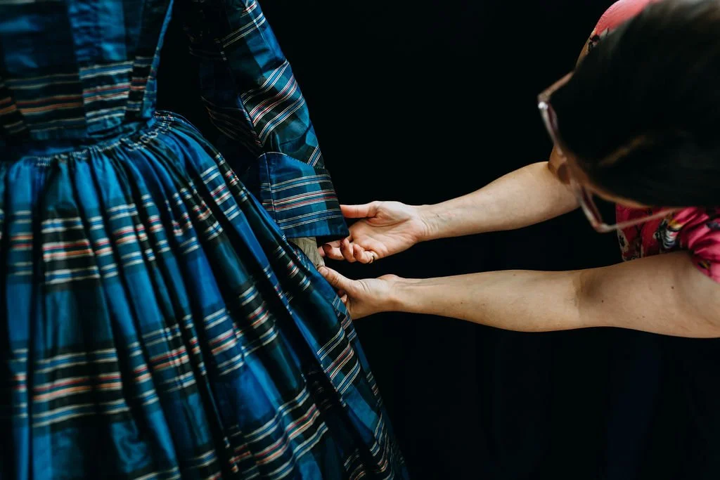 Image shows the waist, upper skirt, and sleeve of blue plaid dress from the late 1850s, with the arms and head of fashion historian Whitney Robertson shown adjusting the sleeve.