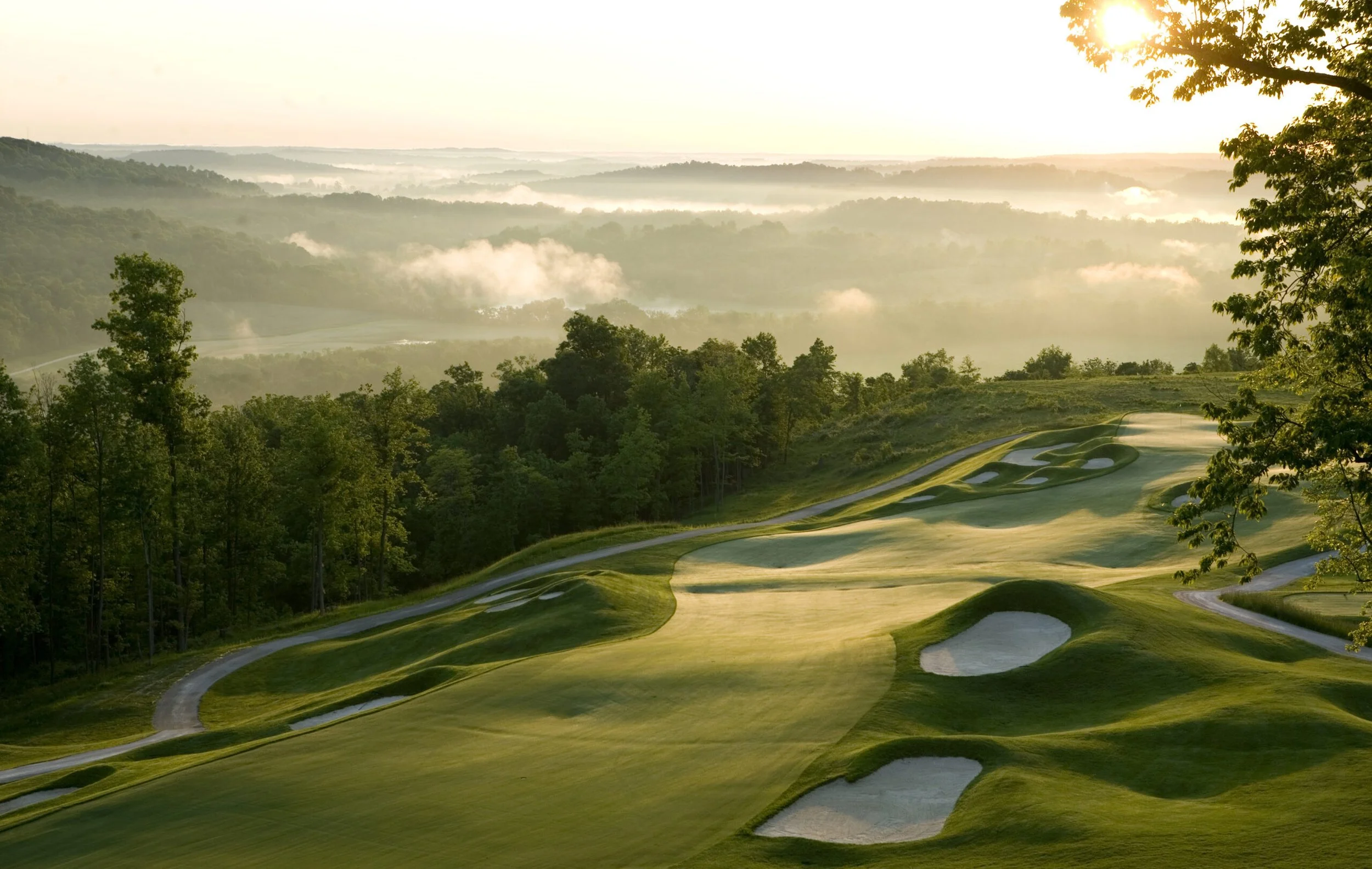 Early morning view of a golf course with sand traps, lush grass, and trees, overlooking a misty valley with rolling hills and a sunrise in the background.
