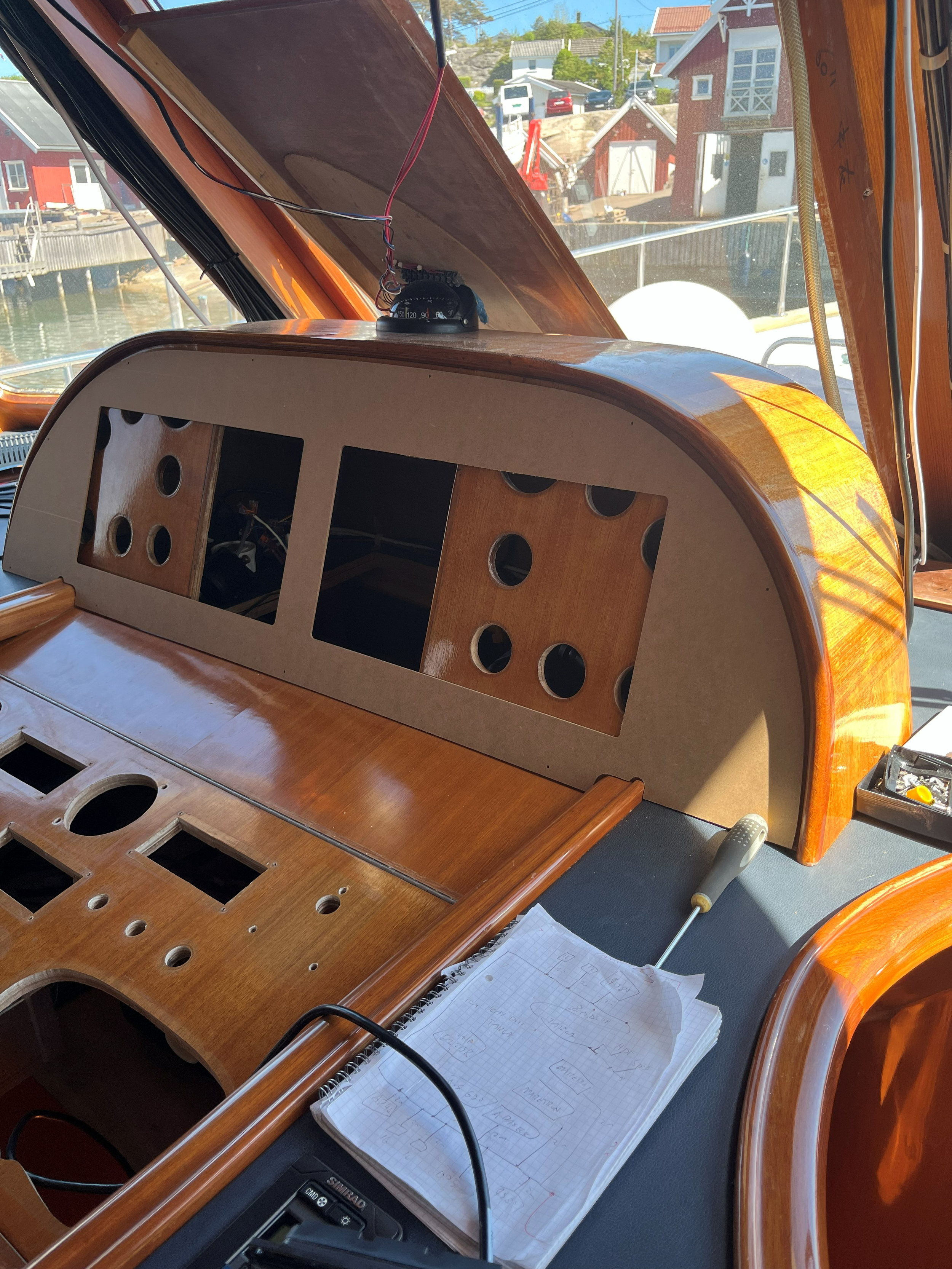 Interiør i en trebåt med styrepanel, kompass, ledninger og kartskisse, sett mot en brygge med tradisjonelle sjøhus.