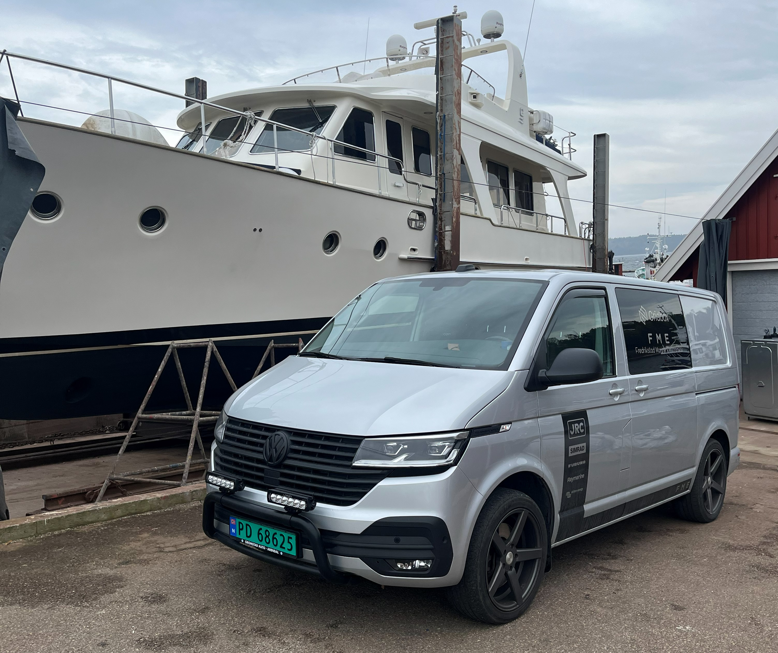 En hvit yacht står på land på Engelsviken Slipp, med Fredrikstad marine elektronikk AS servicebil parkert ved siden av. Flere båter er synlige i bakgrunnen, og det er overskyet vær.