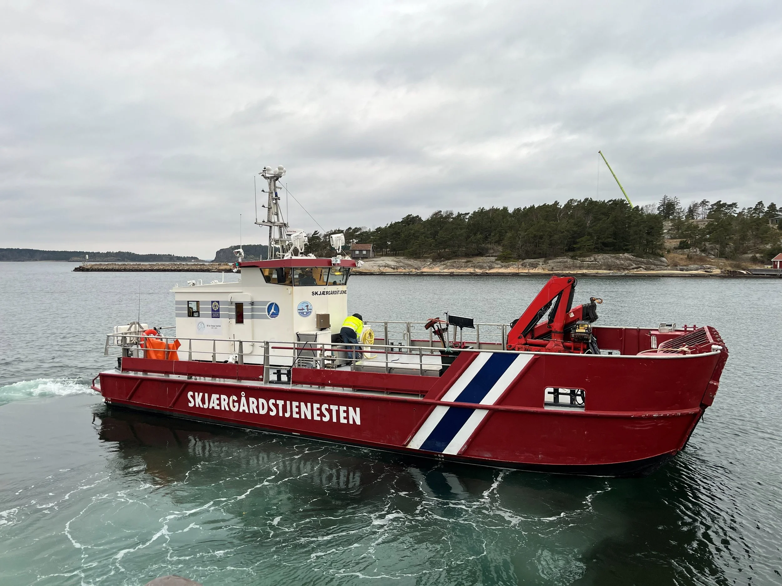 Et yrkesfartøy fra Skjærgårdstjenesten seiler gjennom vann, med en person som jobber på båten. Det er en overskyet dag med skogkledd land i bakgrunnen. Bildet er tatt ved Engelsviken Slipp