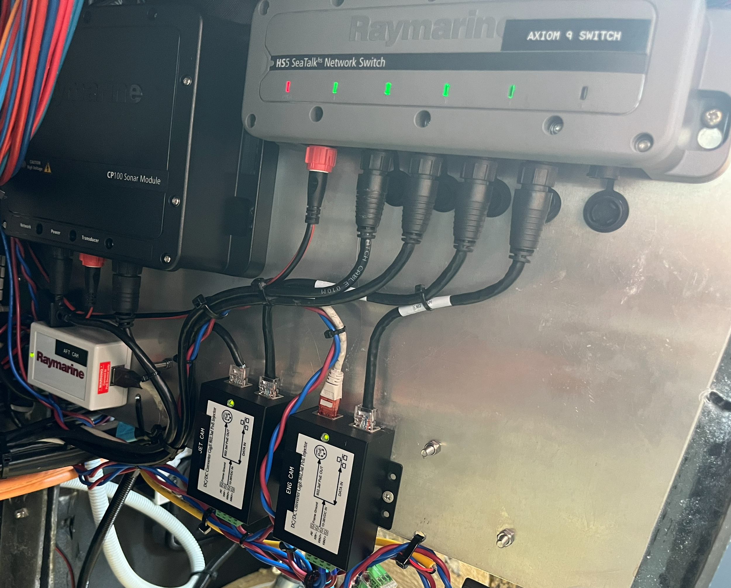 Elektroniske komponenter og kabler, inkludert en Raymarine , en Raymarine HS5 SeaTalkhs nettverkswitch med indikatorlys, og andre koblinger, montert på en vegg.