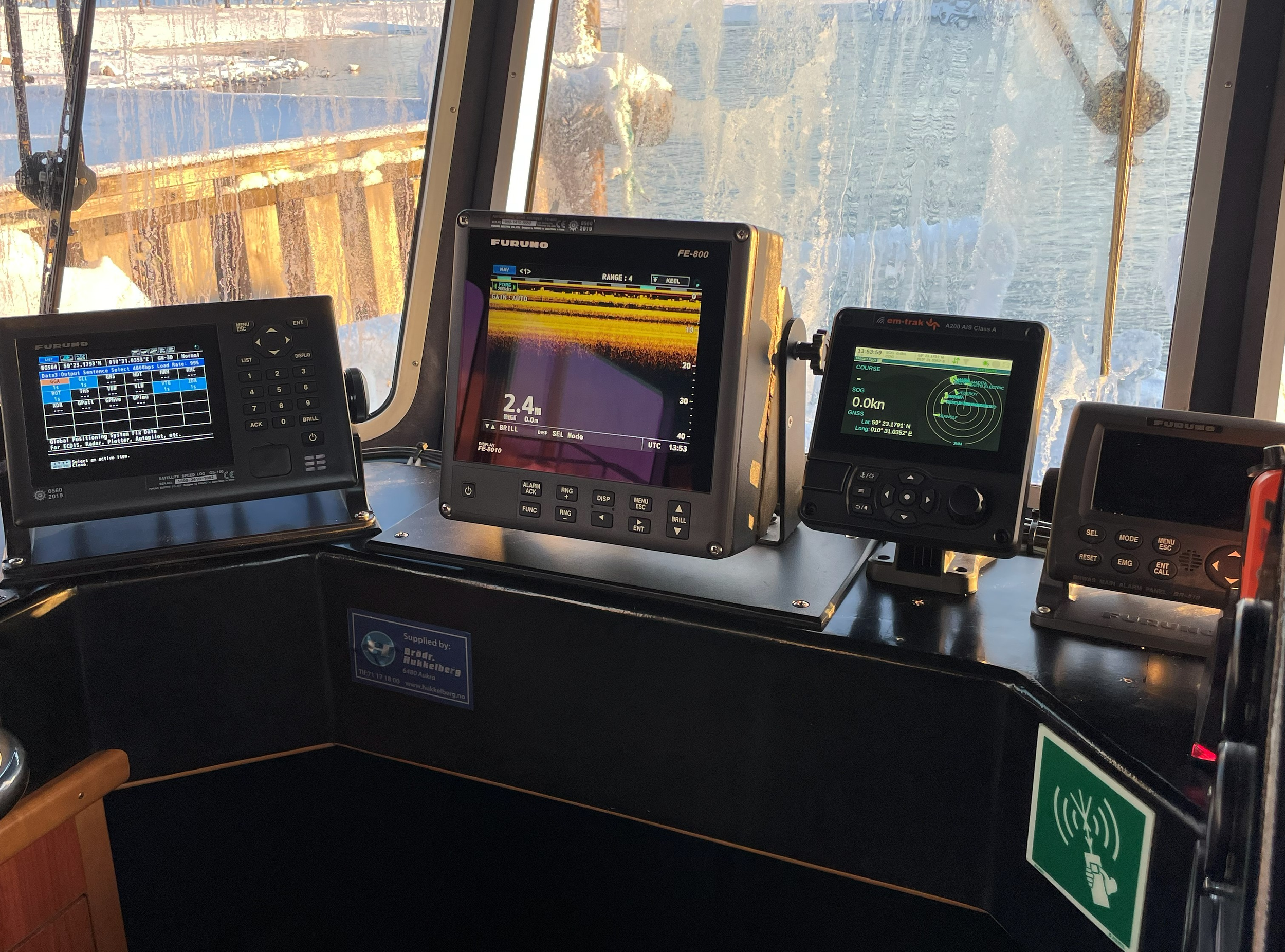 dashbord på yrkesfrtøy med ulike navigasjons- og kommunikasjonspaneler fra Furuno og emtrak ais foran store vinduer som viser snødekte landskap og vann.