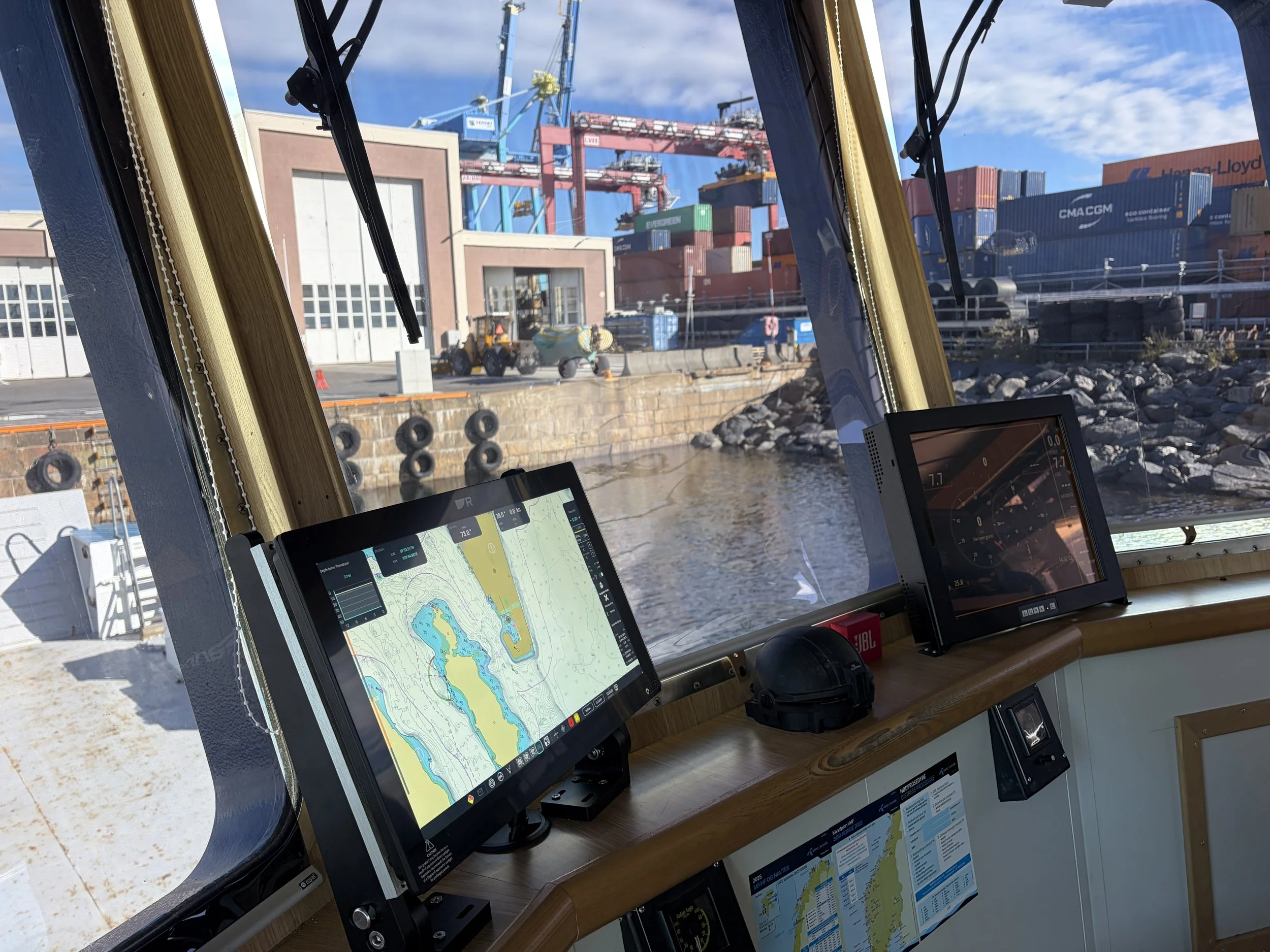 Bildet viser yrkesfartøy arbeidsbåt med raymarine teledyne navigasjonskart på skjermene, med en havn og containerships i bakgrunnen.