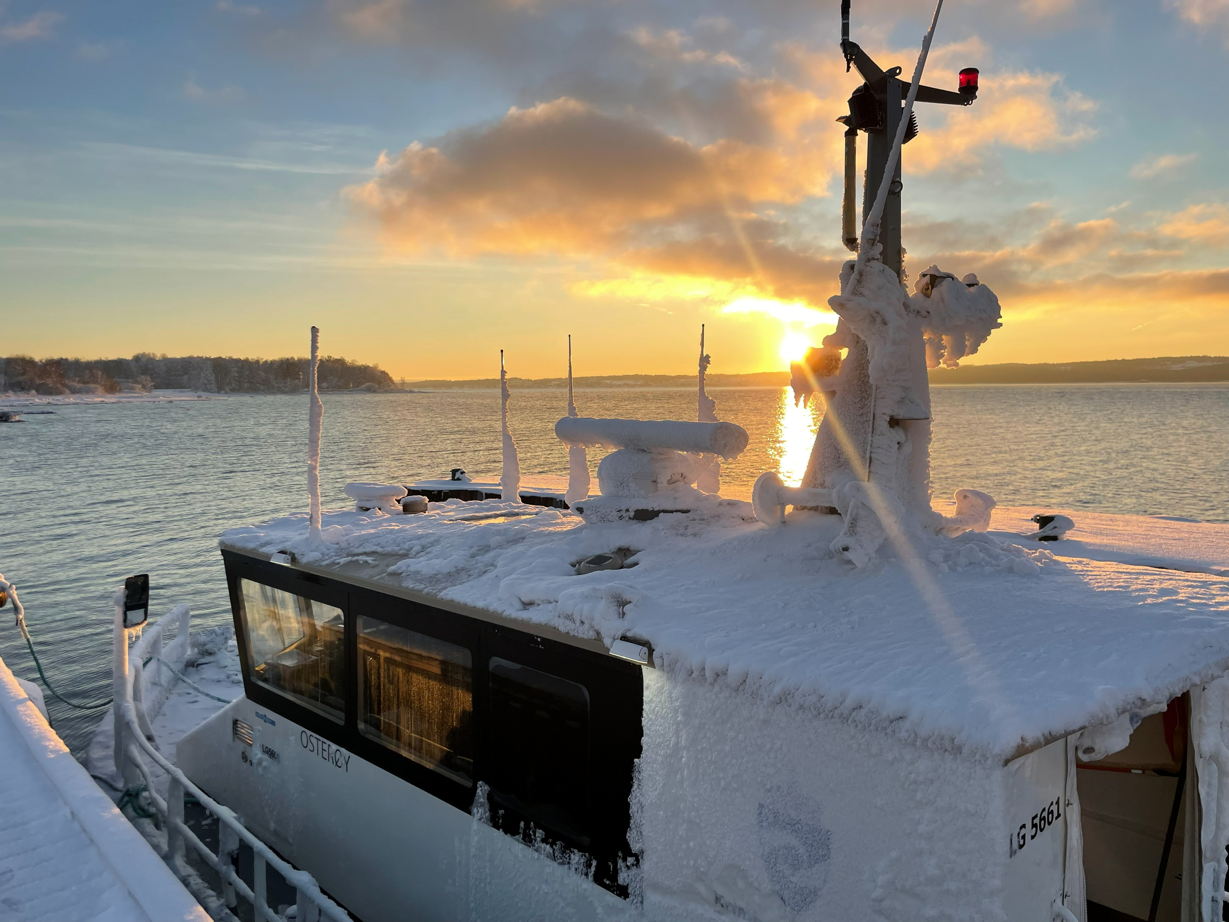 yrkesfartøey er dekket av snø og is, og er plassert ved et vann med en vinterlig solnedgang i bakgrunnen, med skydekke og fjell på horisonten.