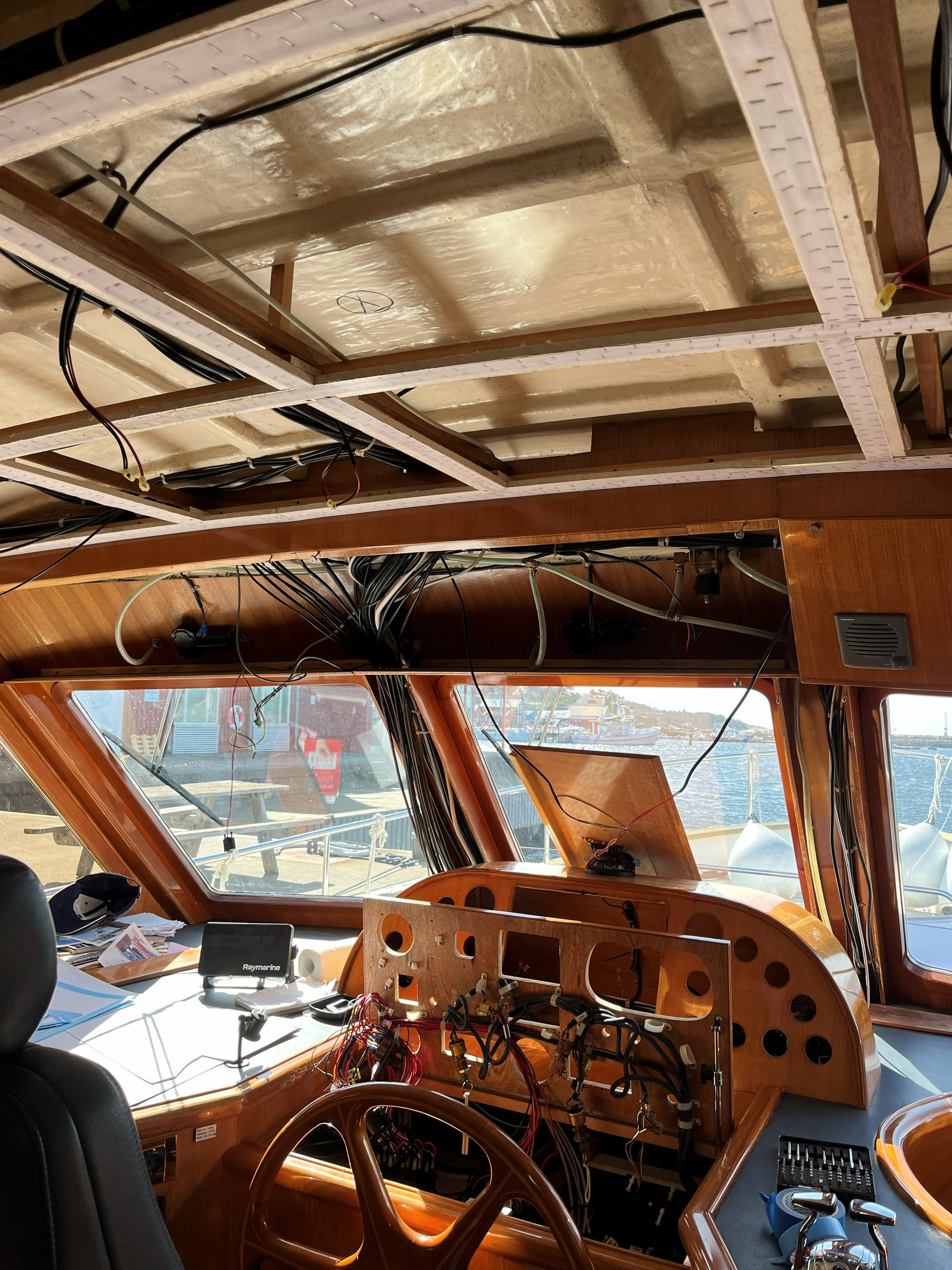 Treinteriør under oppussing med synlige ledninger, instrumentpanel, ratt og utsikt over en marina.