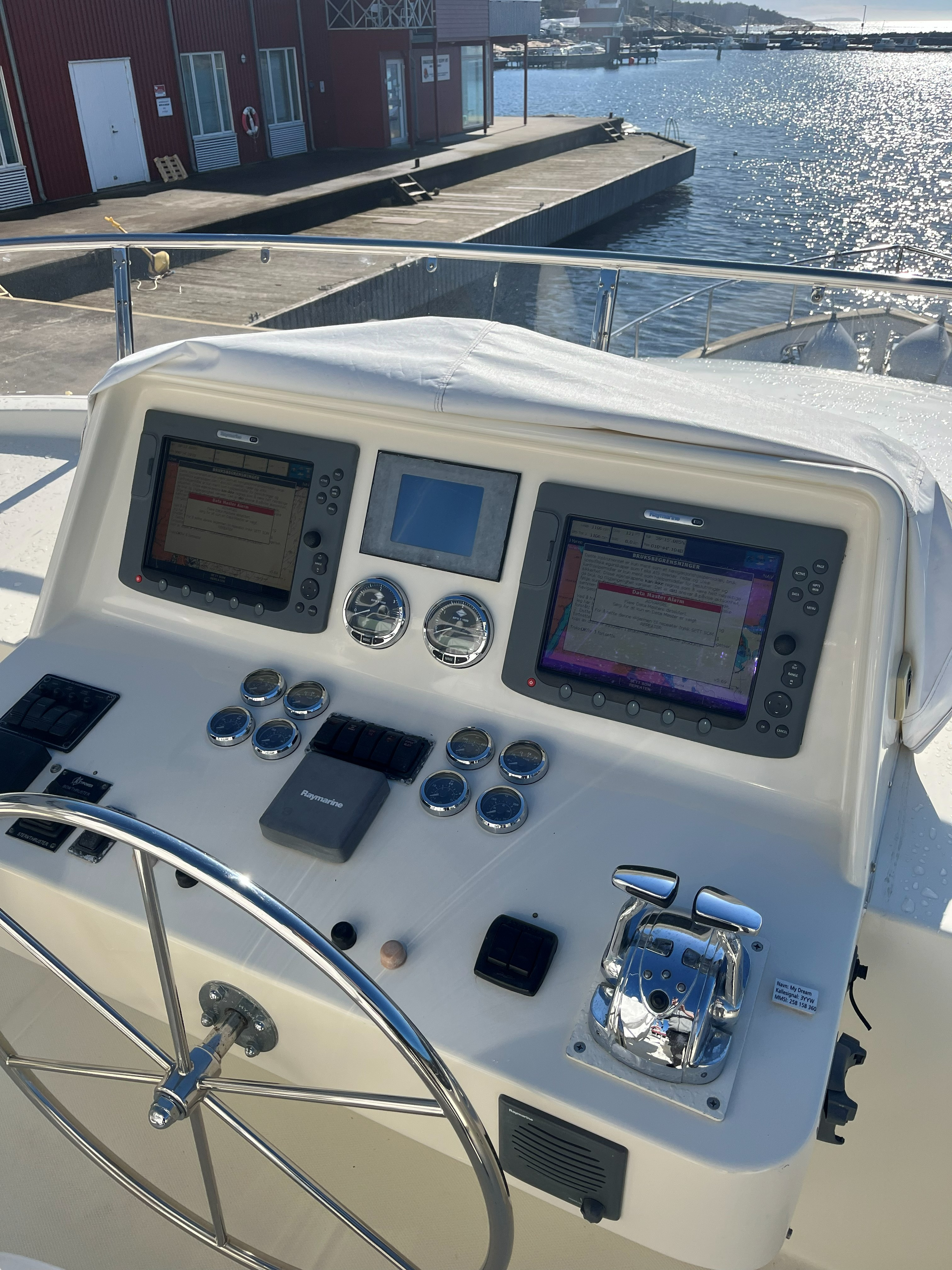 Bildet viser førerposisjonen på en yachten med ulike navigasjonsinstrumenter og styring, med en utsikt over havnen og bryggen i bakgrunnen.