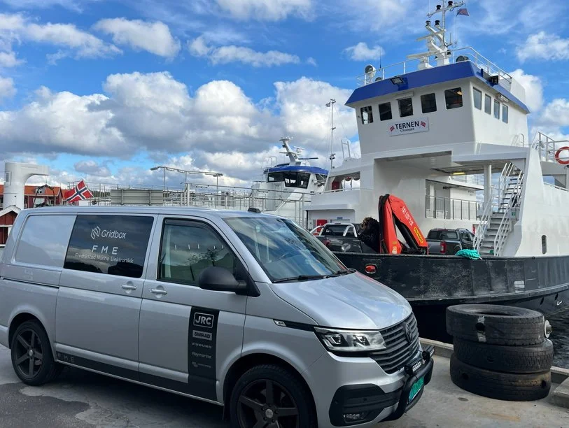 En hvit ferge med flere biler og utstyr på dekk, inkludert et svart og rødt livbåt, og en parkert sølvfarget varebil foran. Det er norske flagg og en blå himmel med skyer i bakgrunnen.