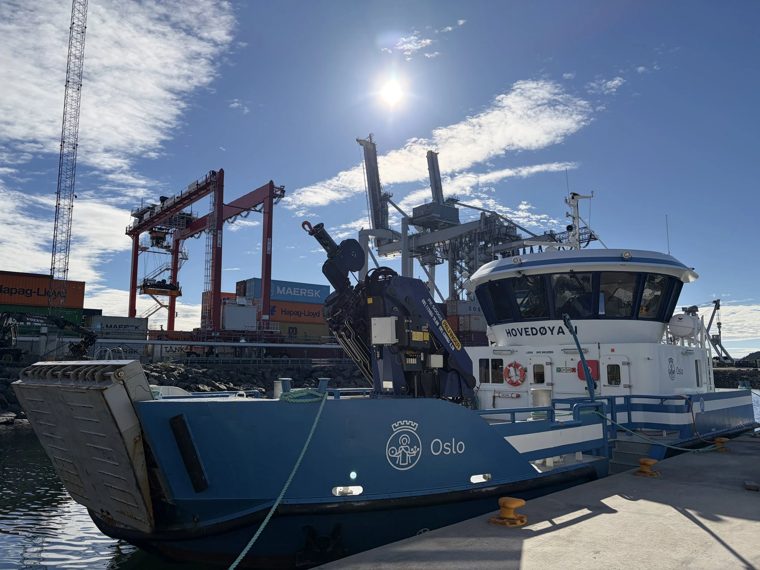 En havn med båter og containerkraner under en solrik himmel med noen spredte skyer. En av båtene har Oslo-logoen på skroget.
