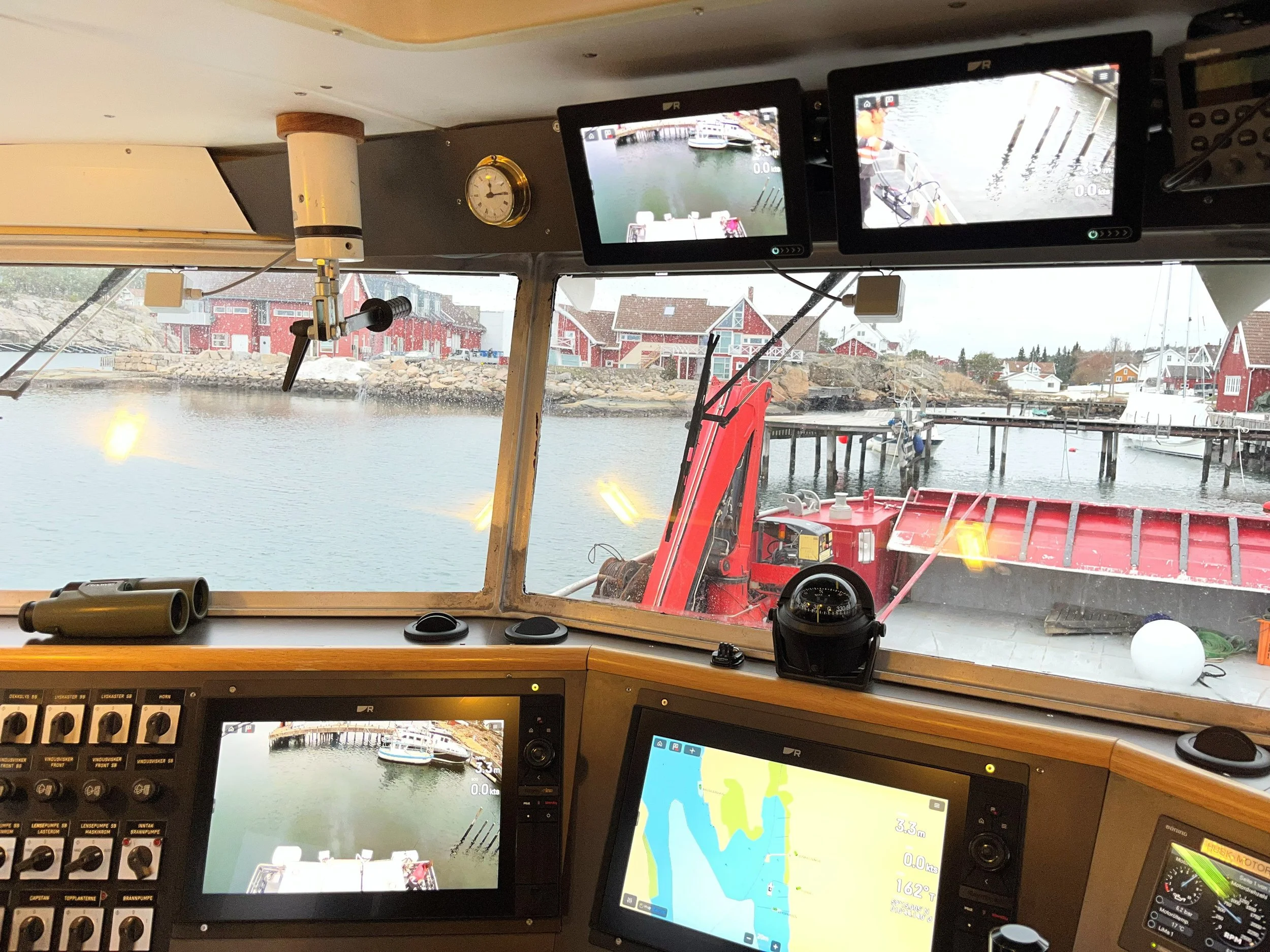 Inne i et yrkesfartøy med flere skjermer og navigasjonsutstyr fra Raymarine, utsikt over en liten havn med båter i Engelsviken og røde bygninger langs vannet.