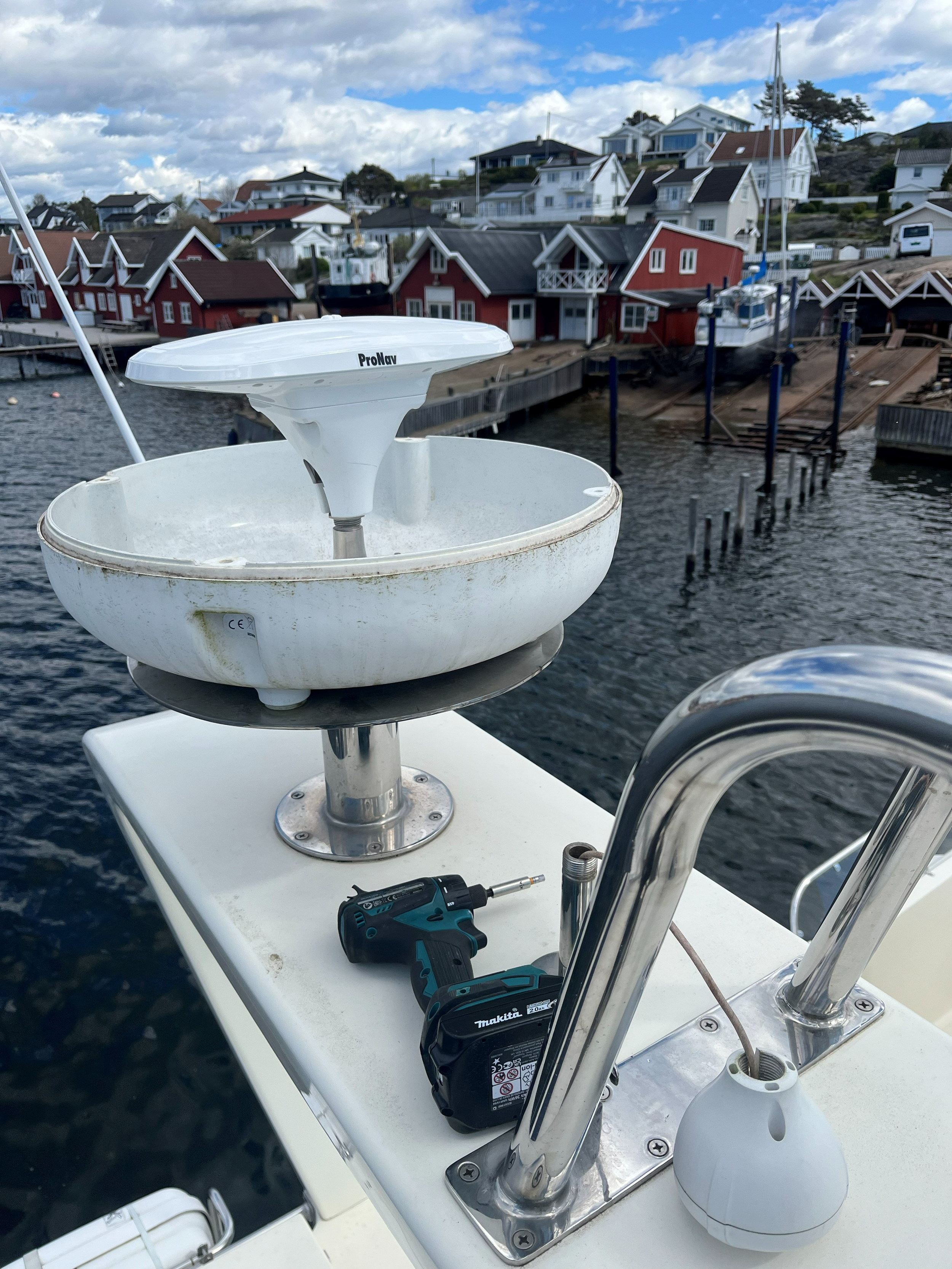 Nærbilde av hvit radar på en båt, med Makita-batteridrill på dekket og rødmalte sjøhus ved havnen i bakgrunnen.
