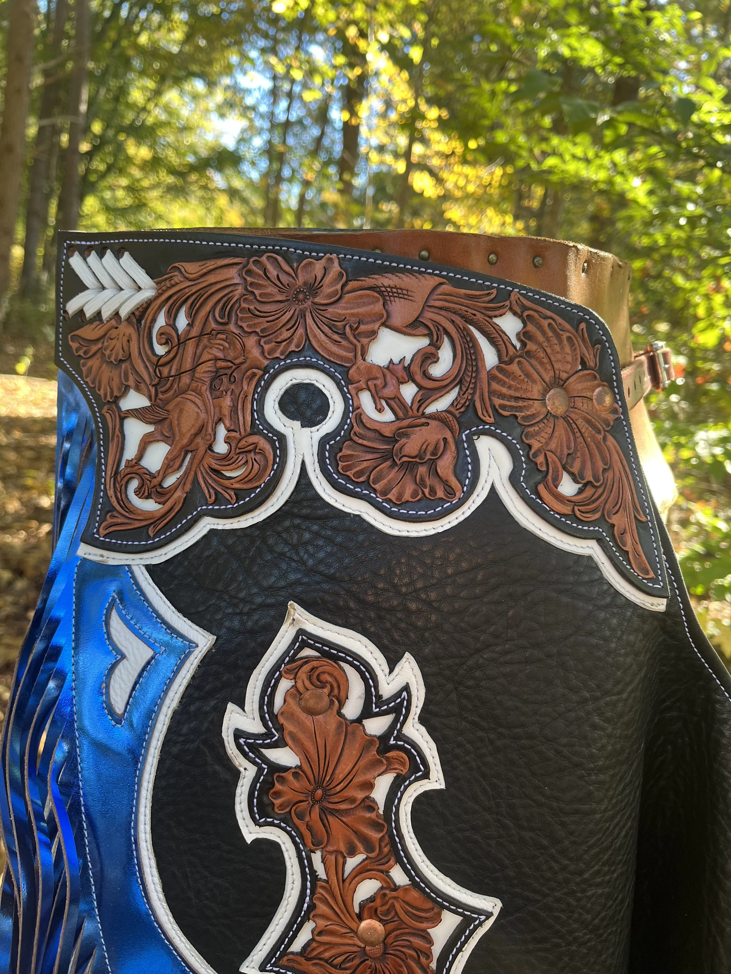 A decorative leather western-style saddlebag with intricate floral and scroll cutout designs, featuring brown, white, and blue accents, hanging outdoors with trees and sunlight in the background.