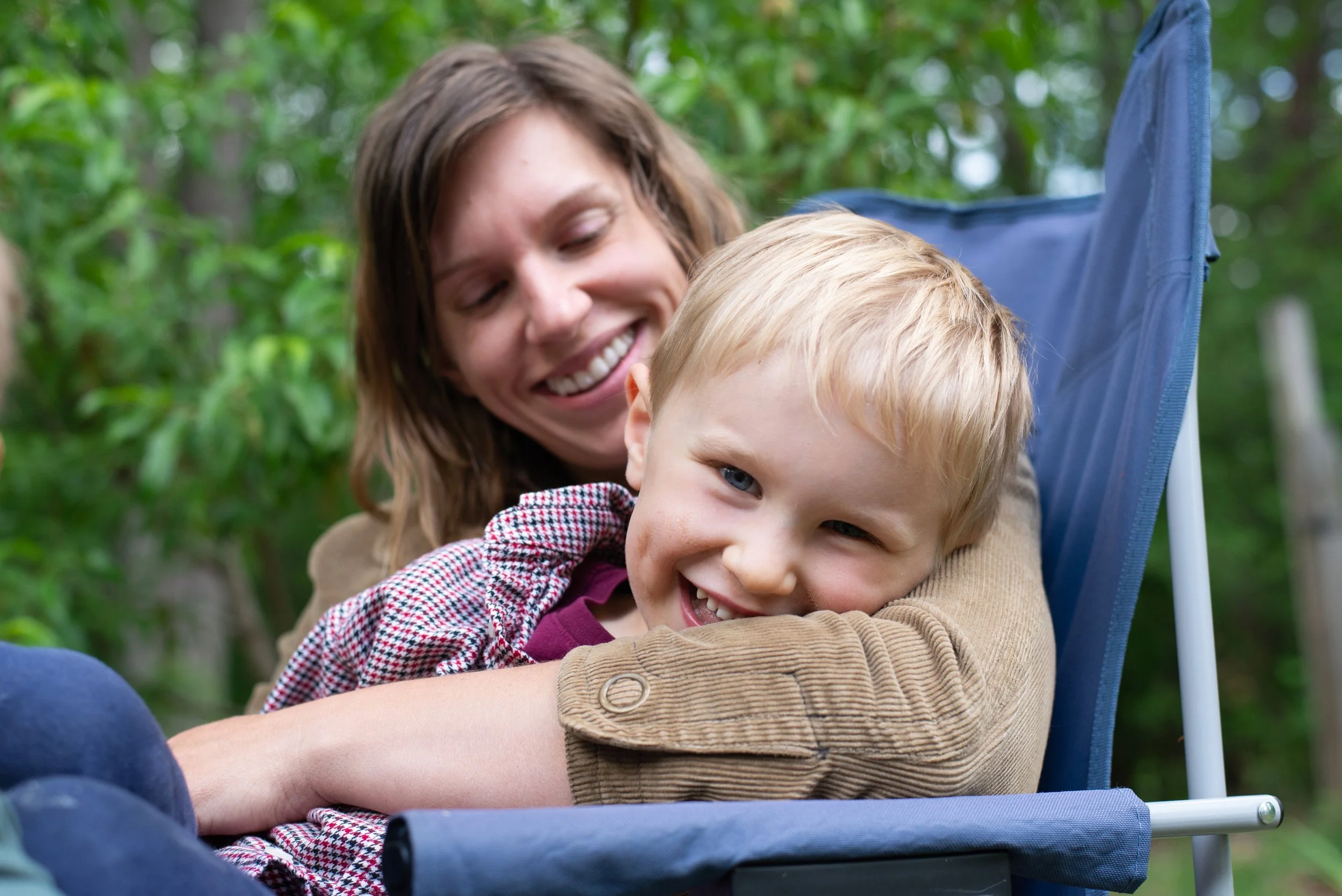 A woman with long brown hair sitting with a young blond boy in a stroller outdoors surrounded by green trees, both smiling and enjoying a moment together.