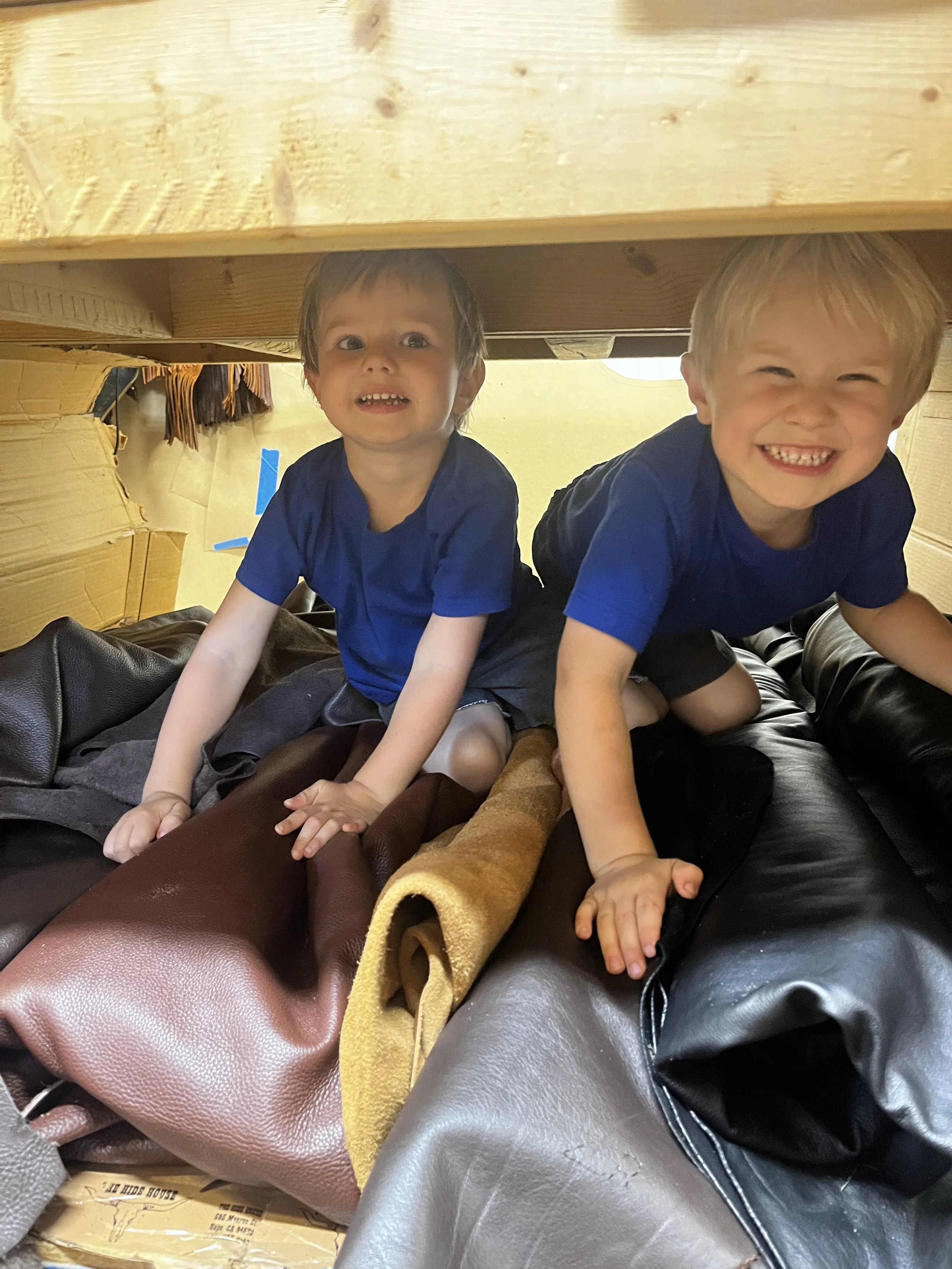 Two young boys crawling under a wooden structure, smiling and looking up at the camera, surrounded by various colored leather sofas.