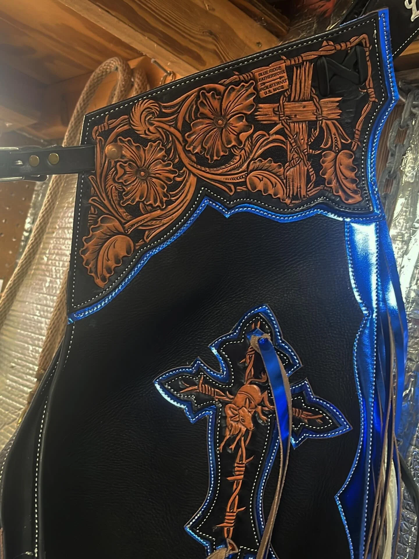 Close-up of a black leather cowboy saddle with ornate floral and cross designs in tan and blue stitching, set against a wooden background.