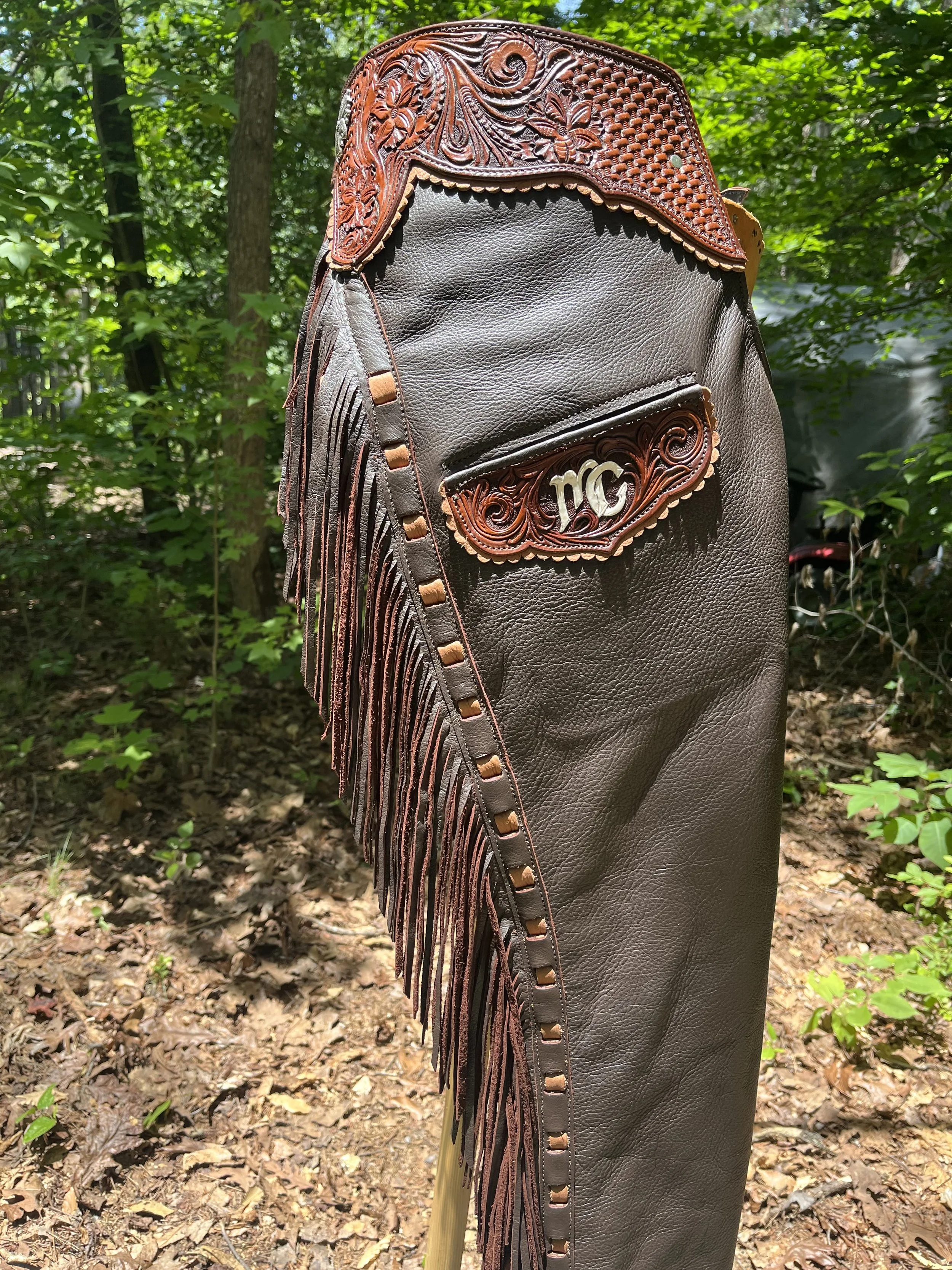 A brown leather western-style guitar strap with detailed tooled leather accents and fringe, mounted on a stand in a wooded outdoor area.