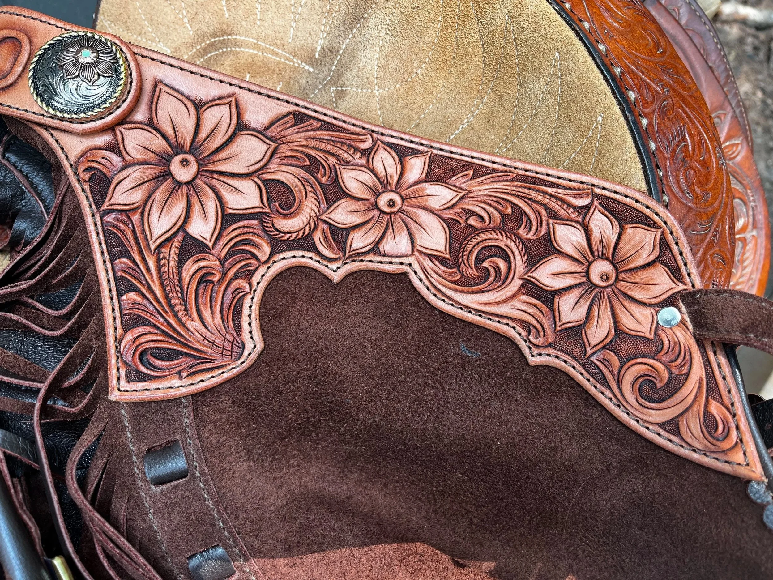 Close-up of a leather saddle with engraved floral patterns and decorative concho, on a textured brown surface.