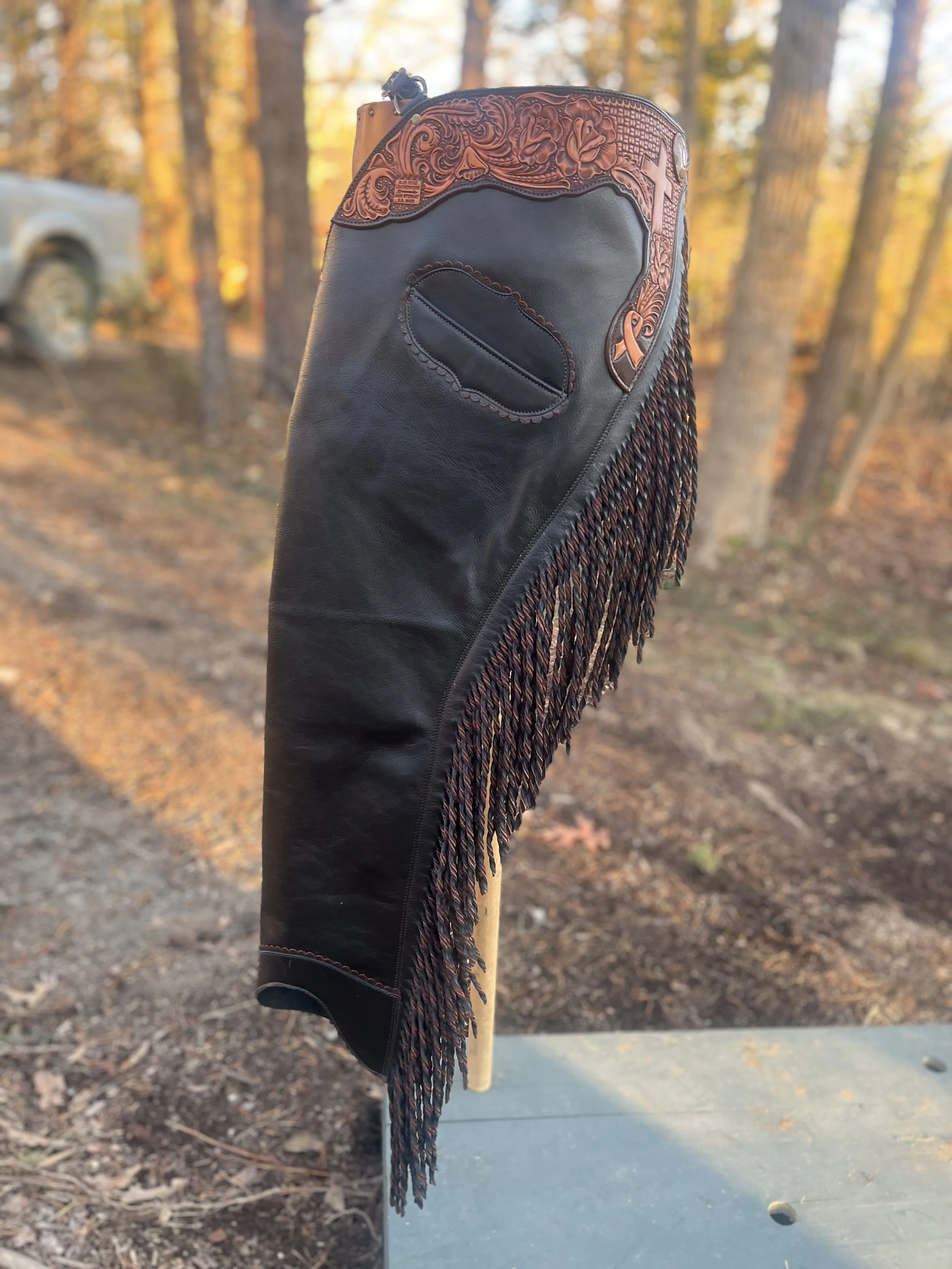 Close-up of a leather golf club headcover with intricate tooled leather at the top and fringe details, standing upright outdoors among trees with autumn foliage.