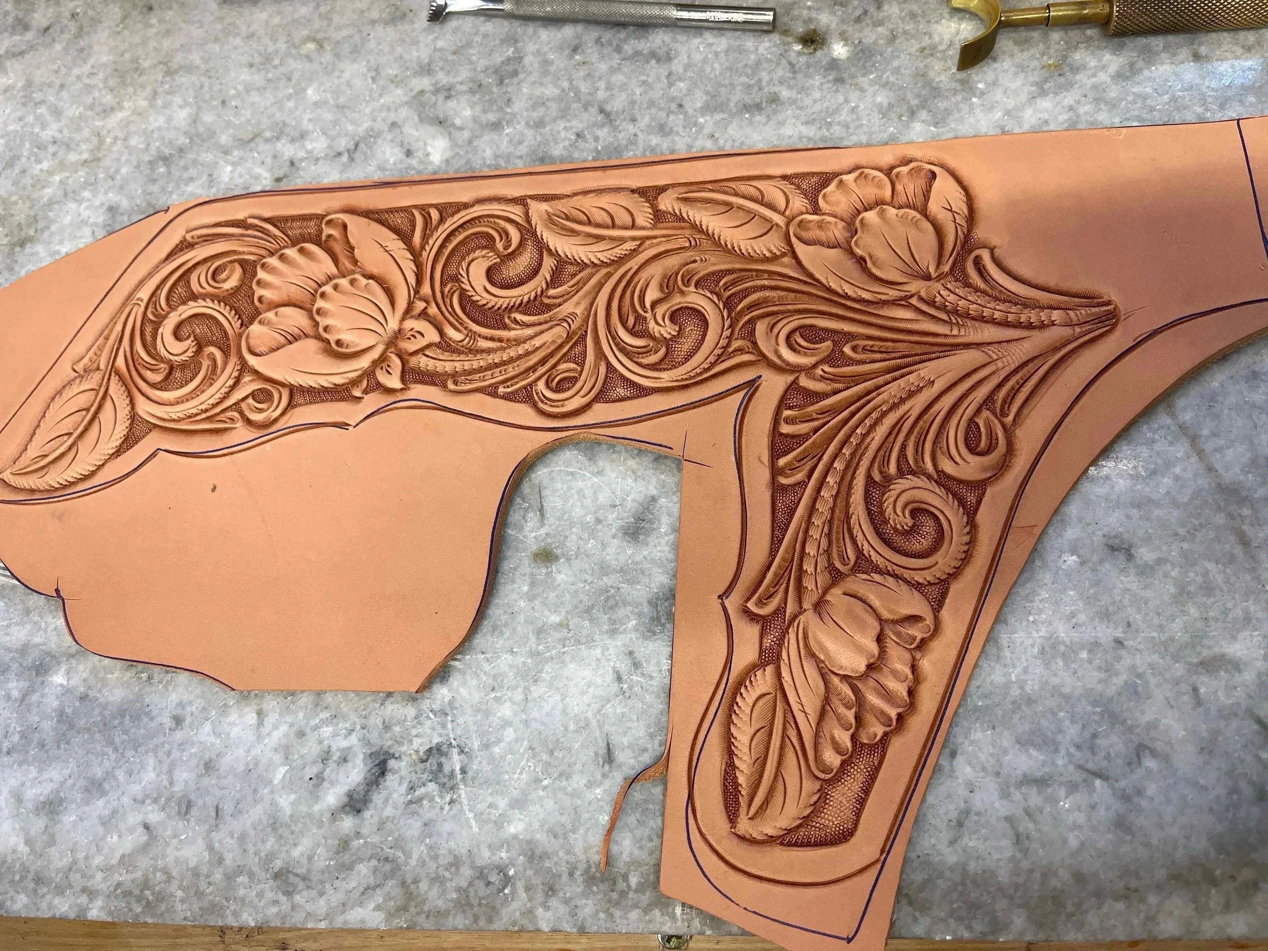 An intricately carved leather piece featuring floral and swirl patterns, placed on a work surface with carving tools visible at the top of the image.