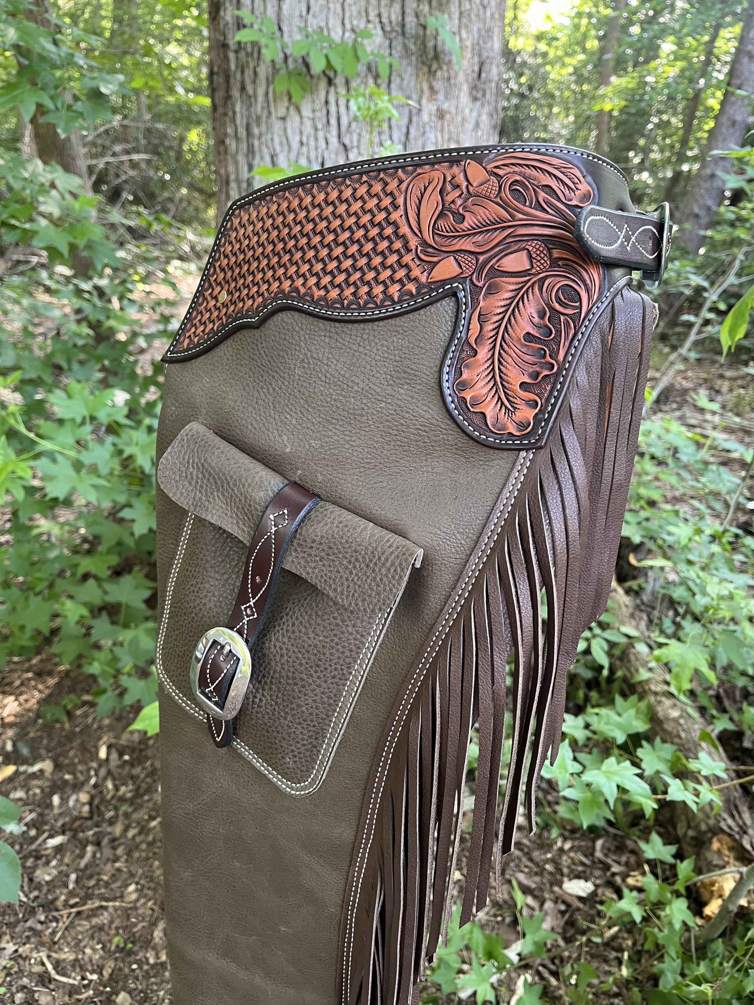 A leather and tooled leather saddlebag with fringe hanging from a post in a wooded area surrounded by green foliage and trees.