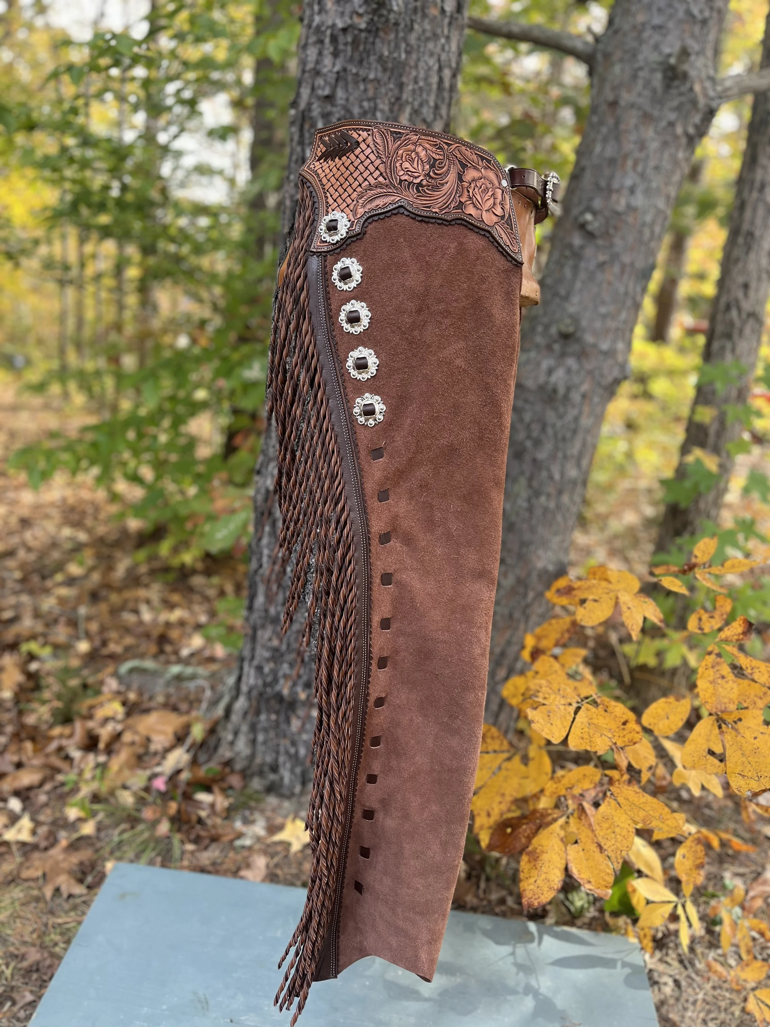 A cowboy boot with floral embroidery, silver conchos, and decorative fringe hanging down the side, set outdoors among orange and green foliage.