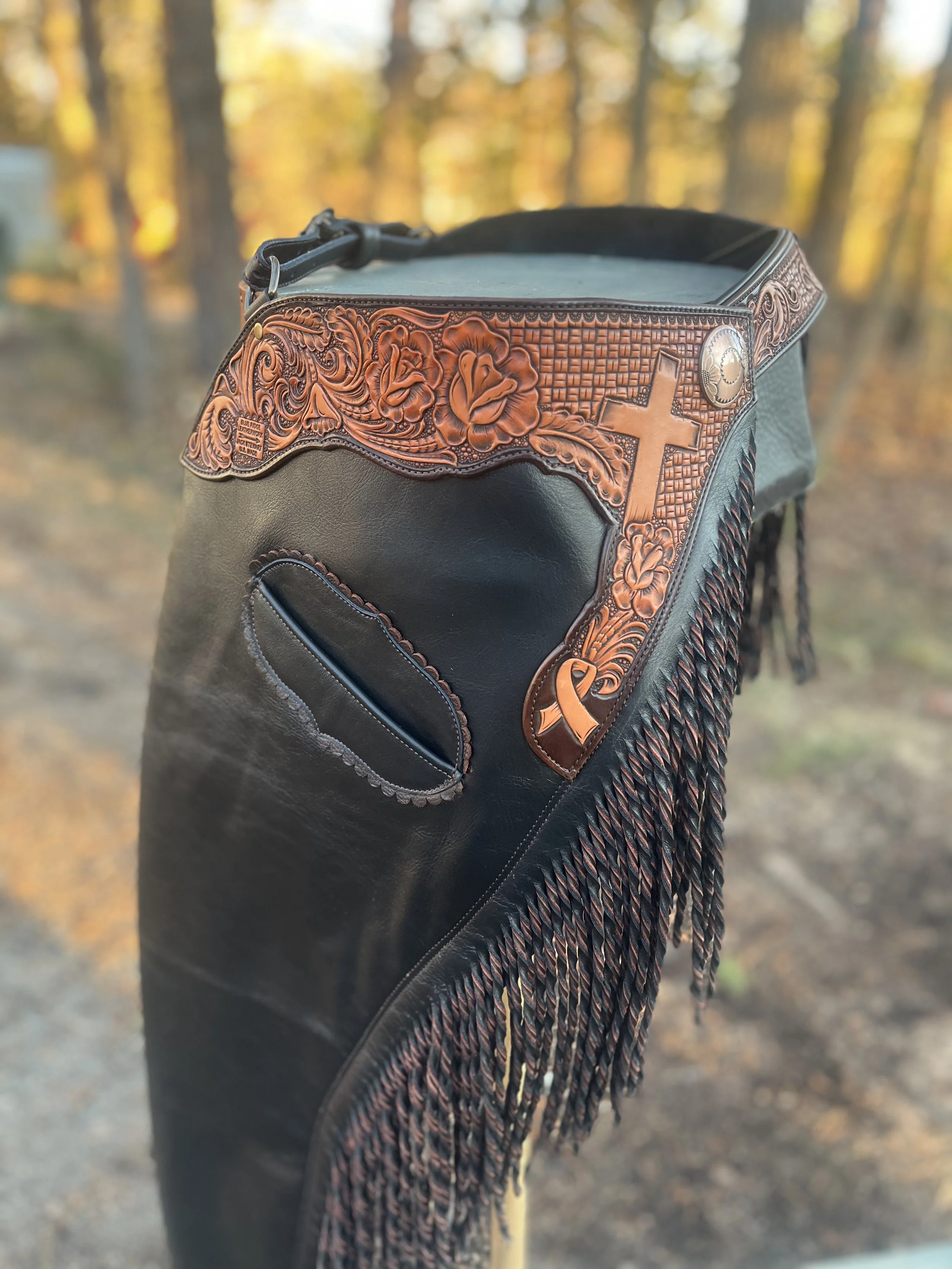 A golf bag with Western-style decorative leatherwork and fringes, set outdoors in a wooded area during autumn.