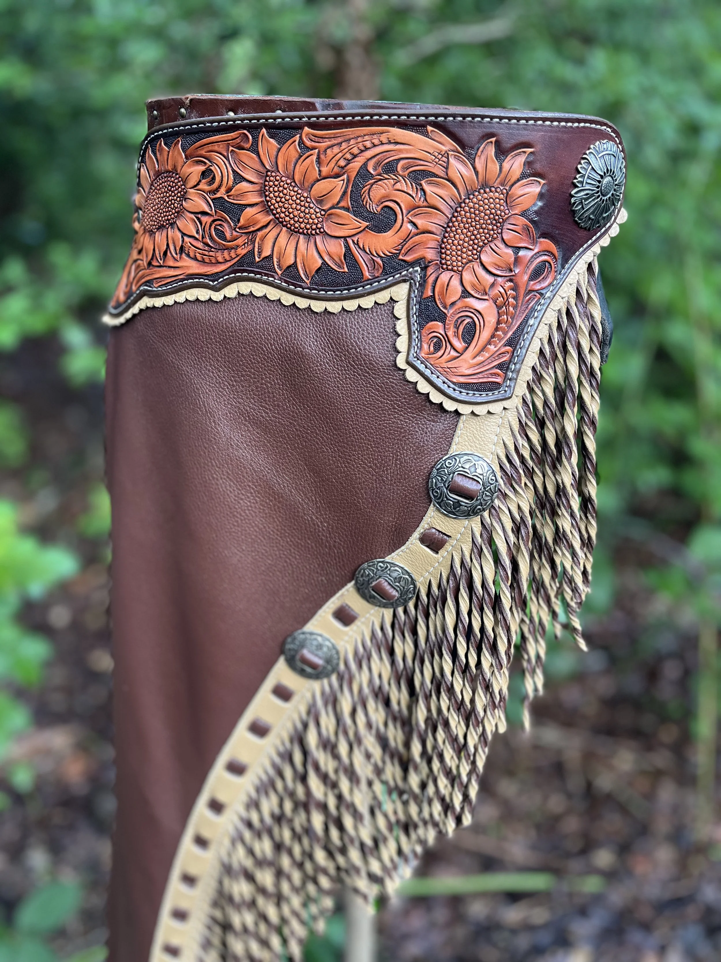 Close-up of a decorative leather cowboy boot with intricate floral and sunflower embossing and metal accents, set against a blurred green outdoor background.