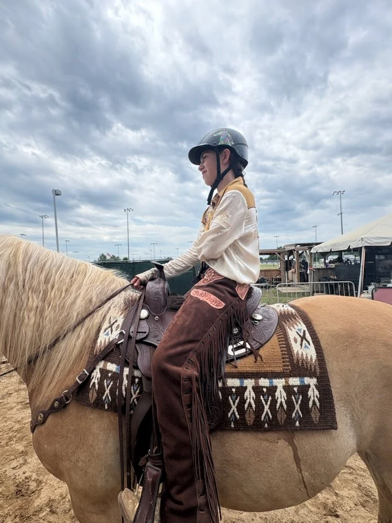A young person wearing a helmet, riding a light-colored horse with a decorative saddle, at an outdoor equestrian event on a cloudy day.