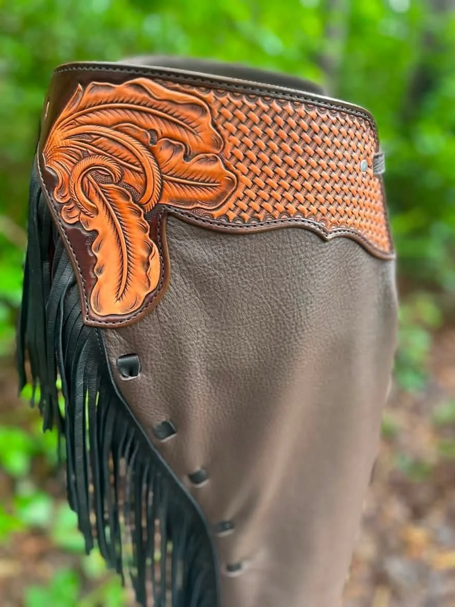 Close-up of a decorative western-style leather saddle or saddle accessory with intricate orange and black leather tooling, fringes, and basket weave pattern on the top, set against a blurred green forest background.