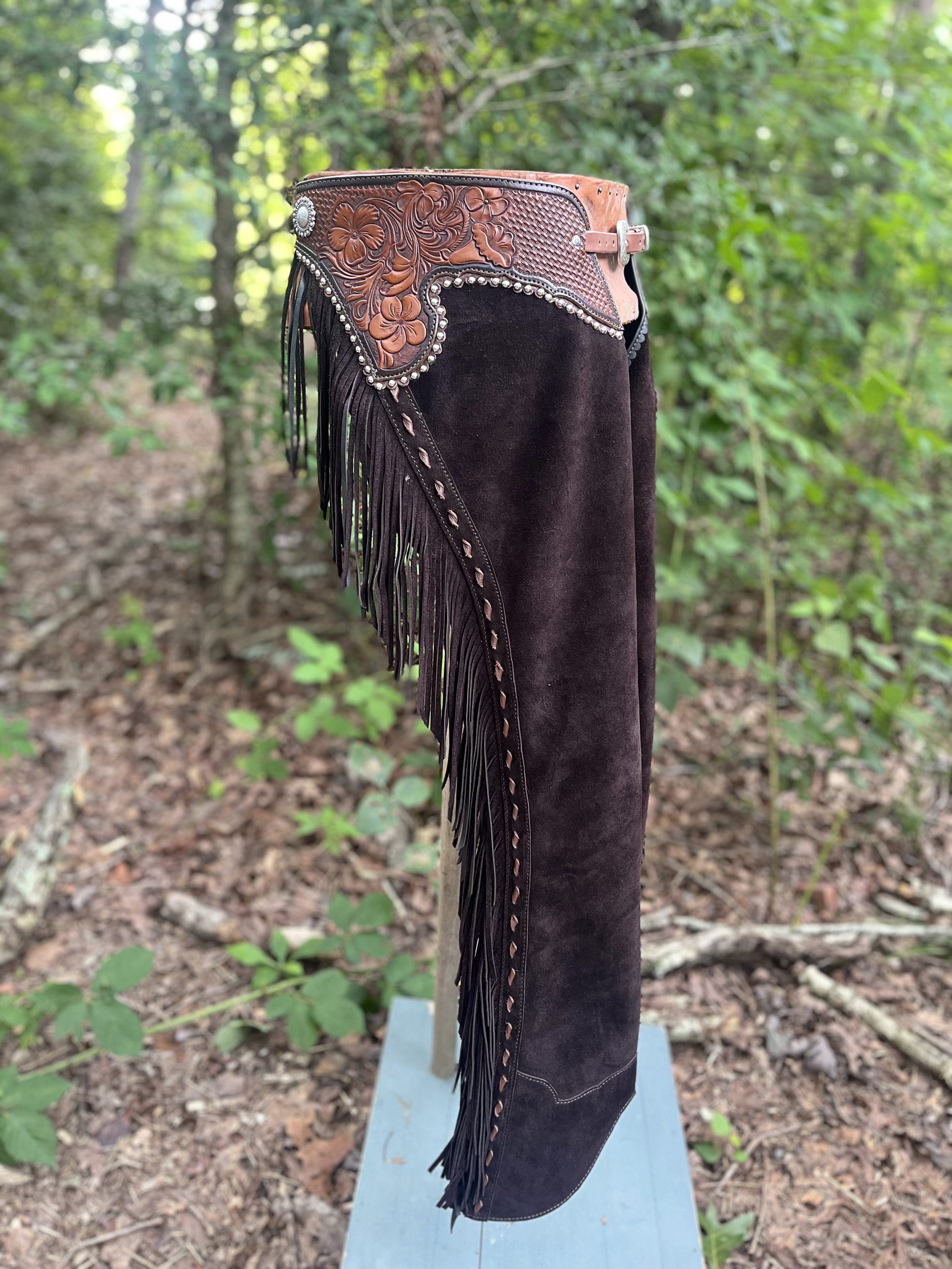 A Western-style cowboy chaps displayed on a blue stand in a forest setting with green foliage and brown ground.