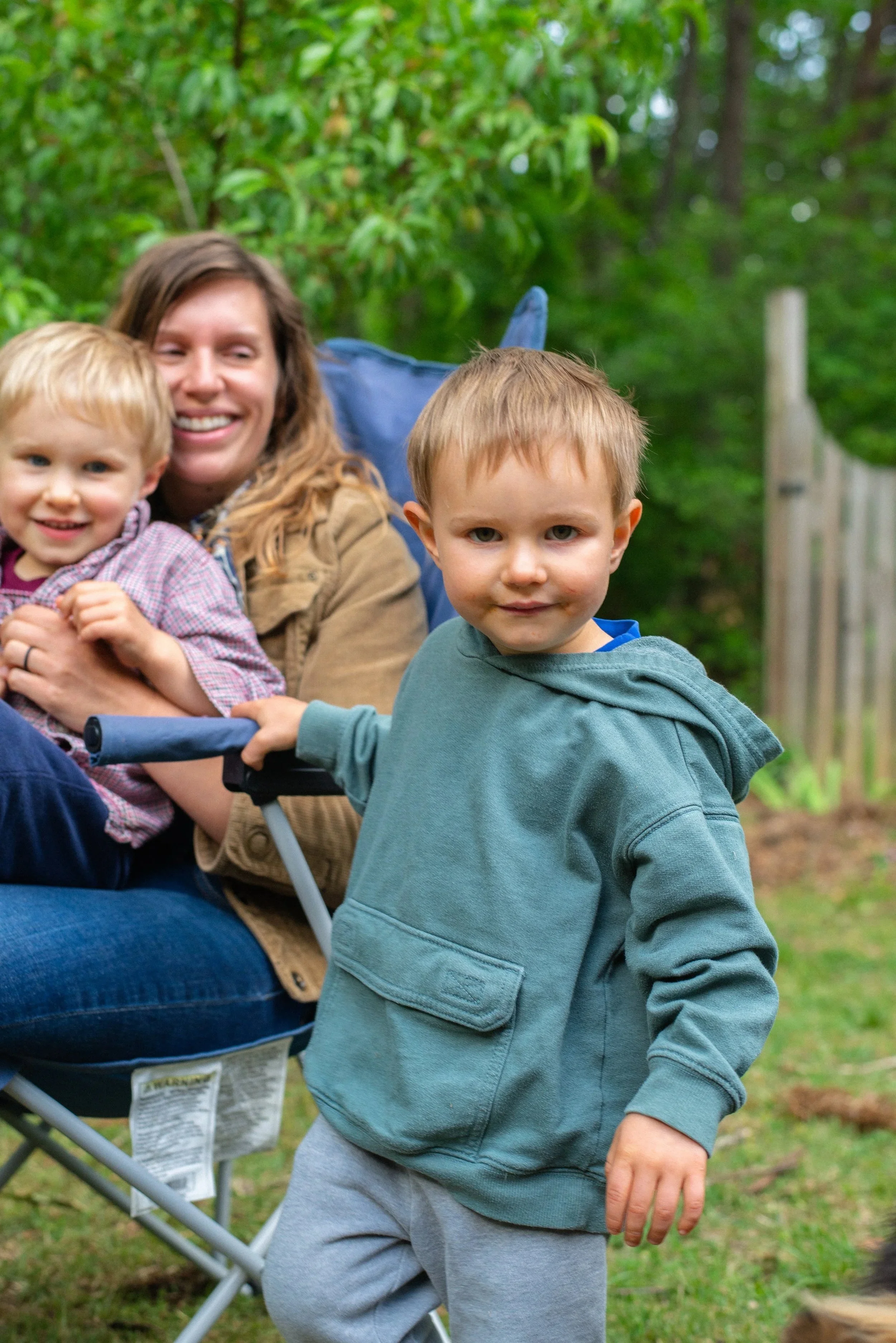 A woman smiling with two young children outdoors in a wooded area. One child is sitting in a stroller, and the other is standing nearby.