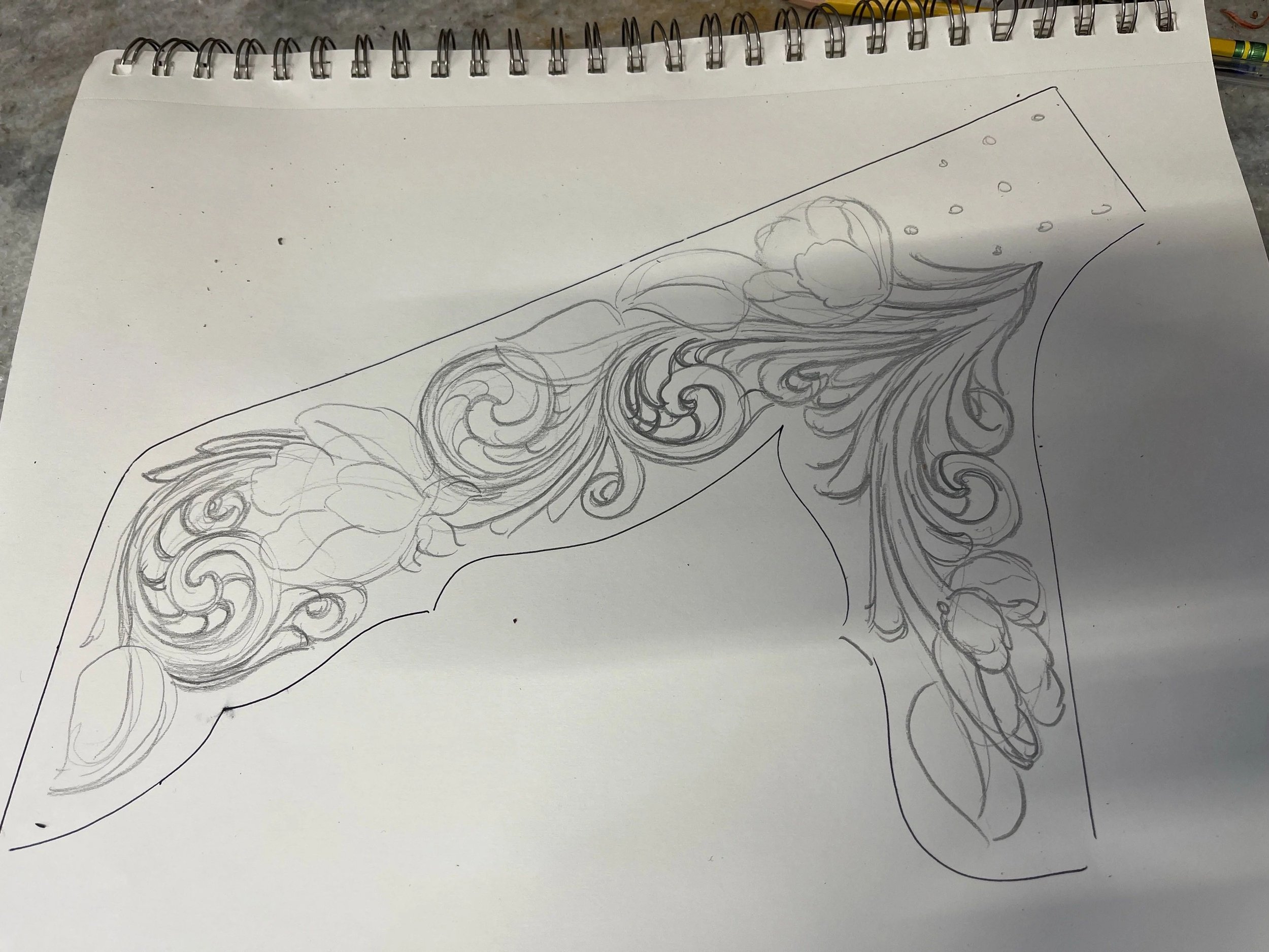 Sketch of floral design on a spiral-bound notebook resting on a gray surface, with a green and yellow pencil visible in the background.