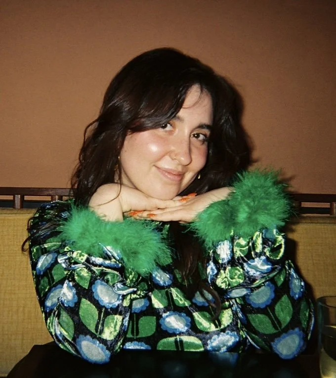A woman with dark wavy hair, wearing a colorful patterned top with green fur cuffs, smiling and resting her chin on her hands, in an indoor setting.