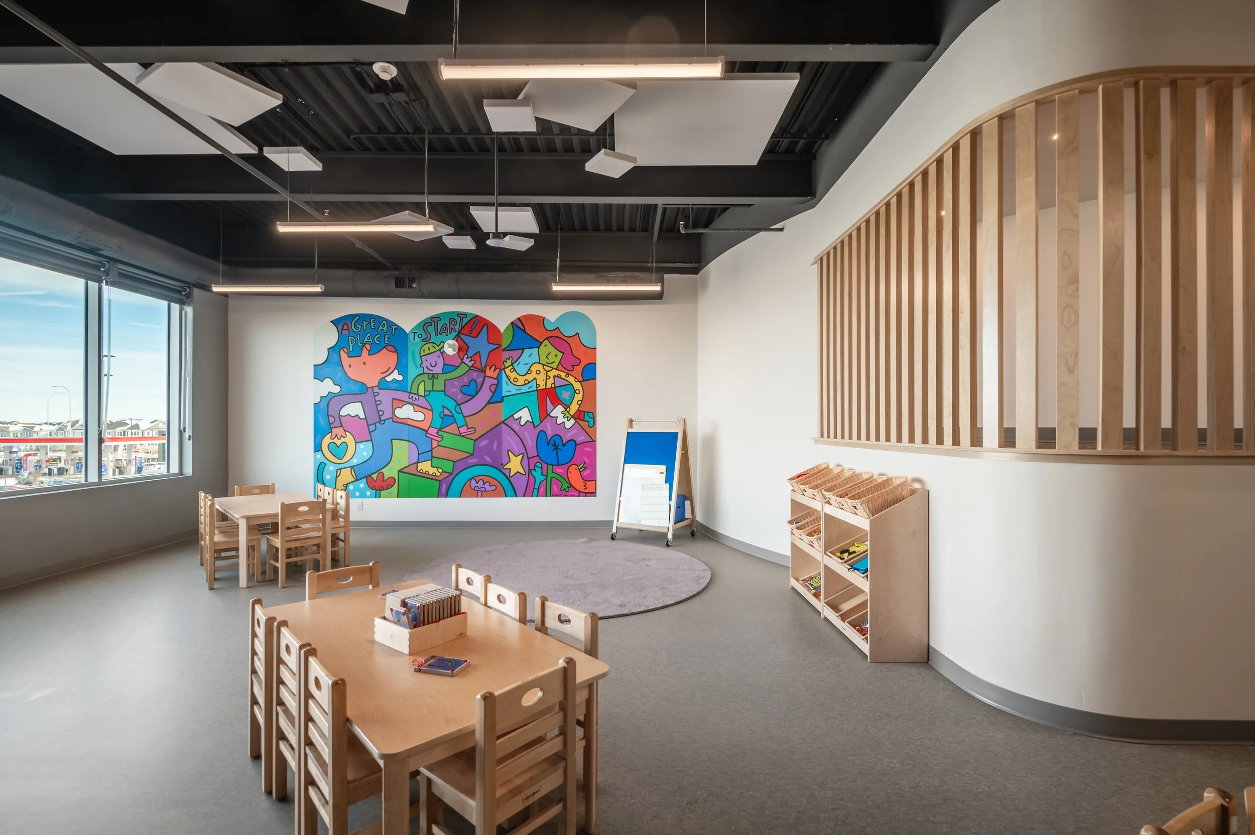 Empty indoor children's play area with wooden tables and chairs, a colorful mural on the wall saying ''A Great Place To Start,'' large windows showing a parking lot outside, and a wooden partition with cubbies.