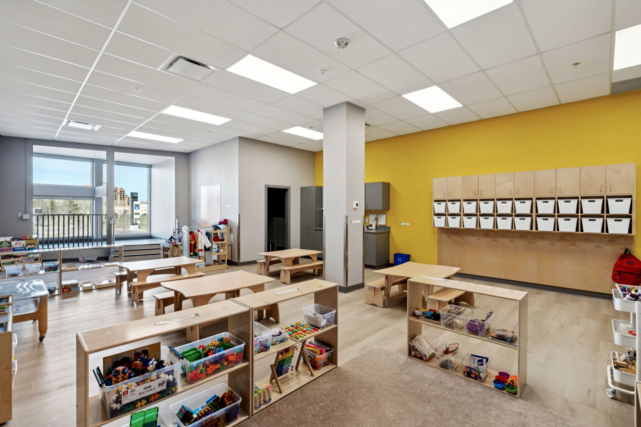 Empty indoor children's play area with wooden tables and chairs, a colorful mural on the wall saying ''A Great Place To Start,'' large windows showing a parking lot outside, and a wooden partition with cubbies.