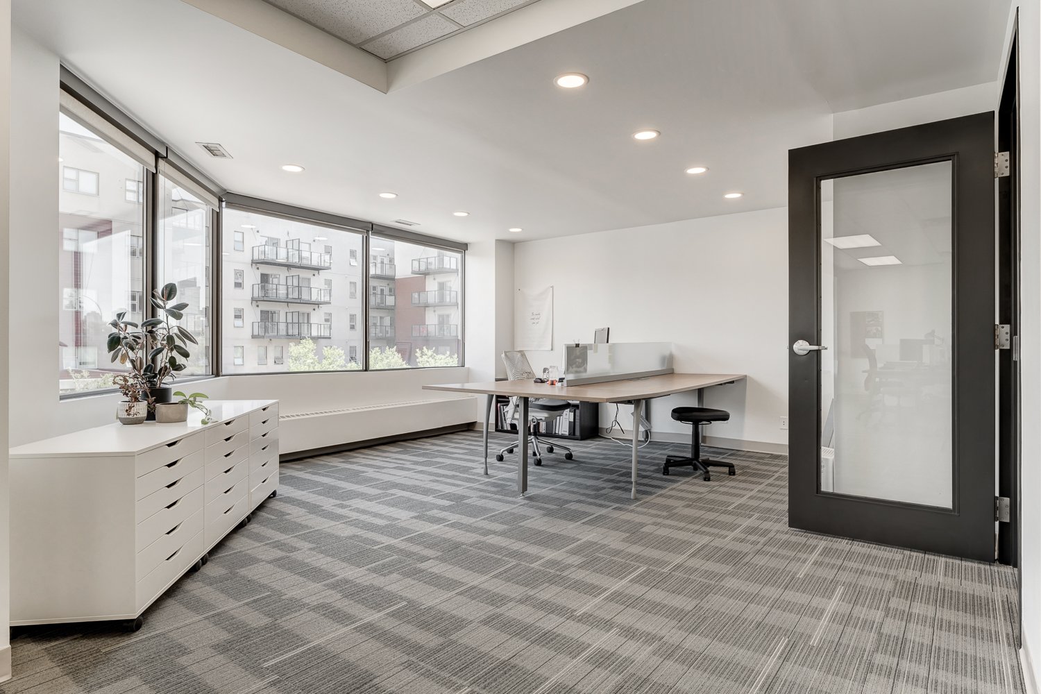 Empty modern office with large windows, a white cabinet with plants, a desk with chairs, and glass door.