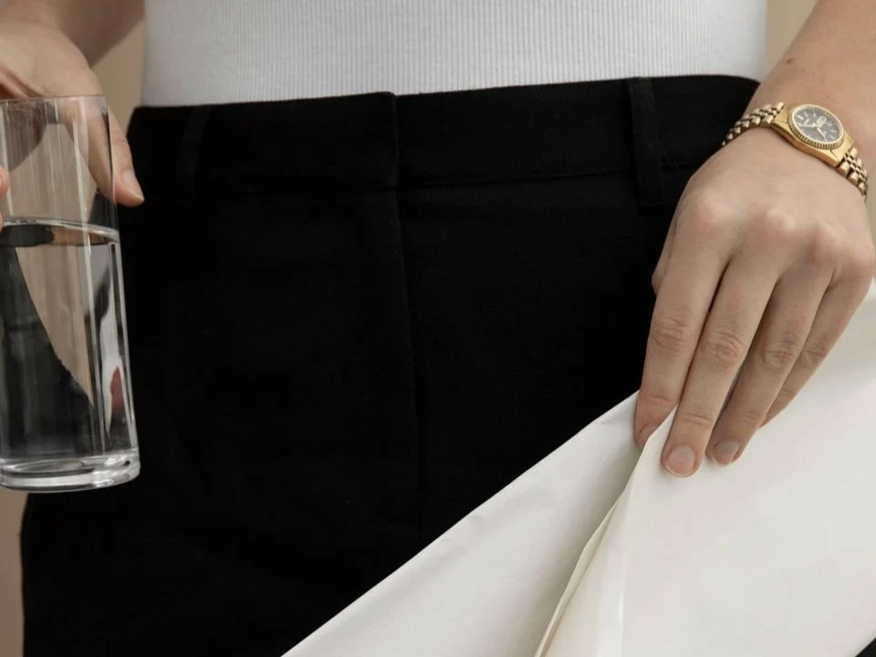 A person holds a glass of water and folded paper while wearing a white top, black trousers, and a gold watch.