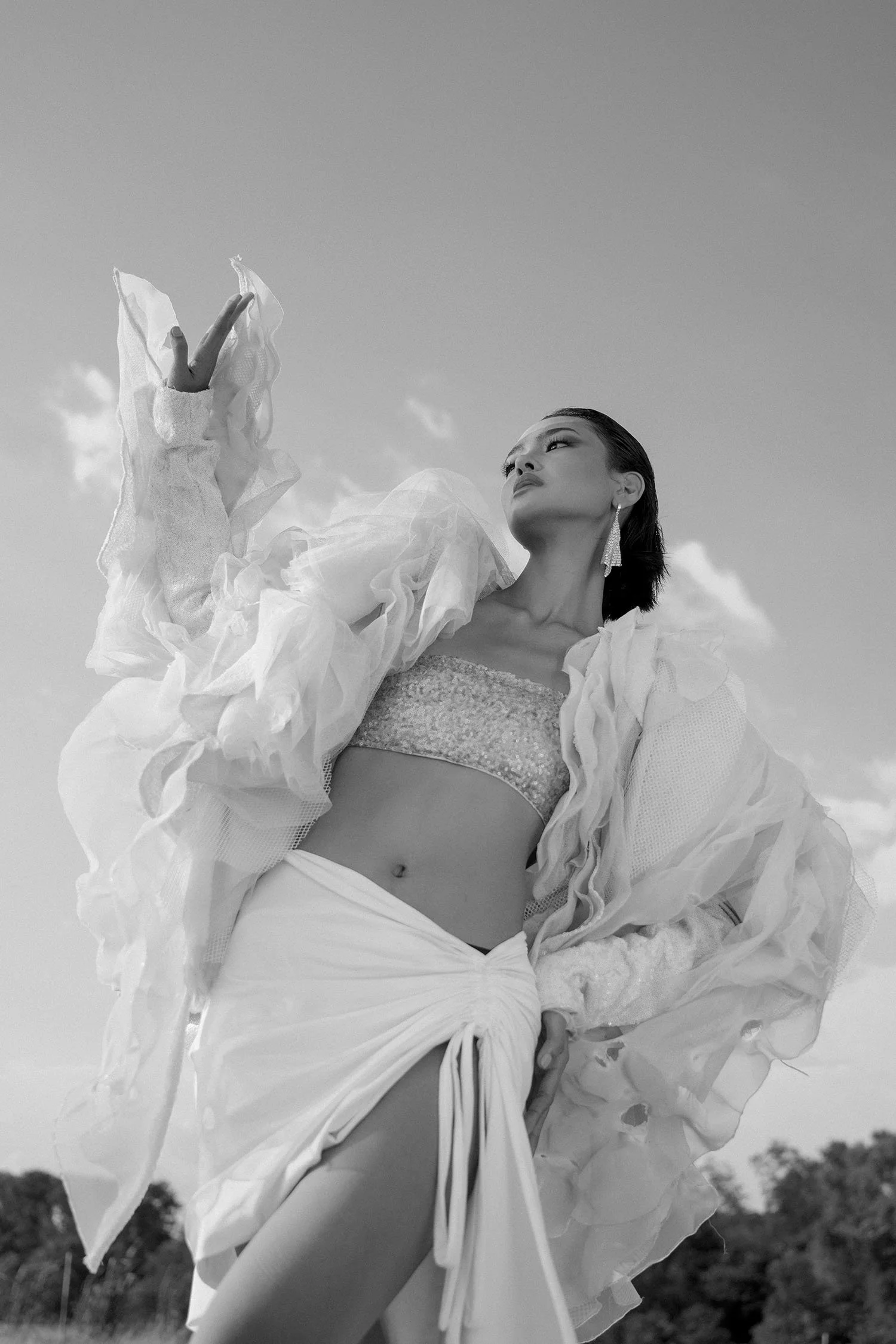 A woman dressed in elaborate, flowing clothing, looking into the distance, standing outdoors against a cloudy sky.
