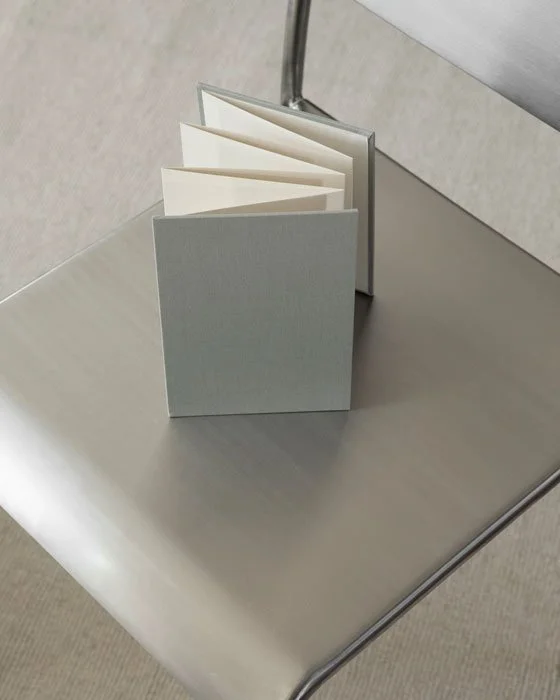 A small, closed, white notebook with blank pages, standing upright on a metallic chair, next to a clear glass or acrylic divider.