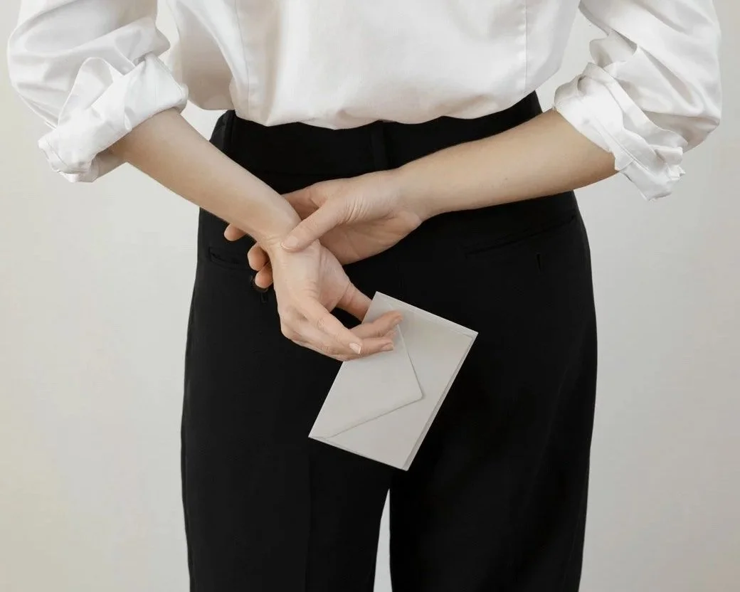 A person dressed in a white shirt and black pants holding a small white envelope behind their back.