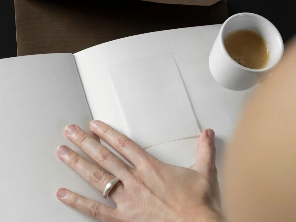 A hand rests on the pages of a blank notebook beside a white cup of espresso.