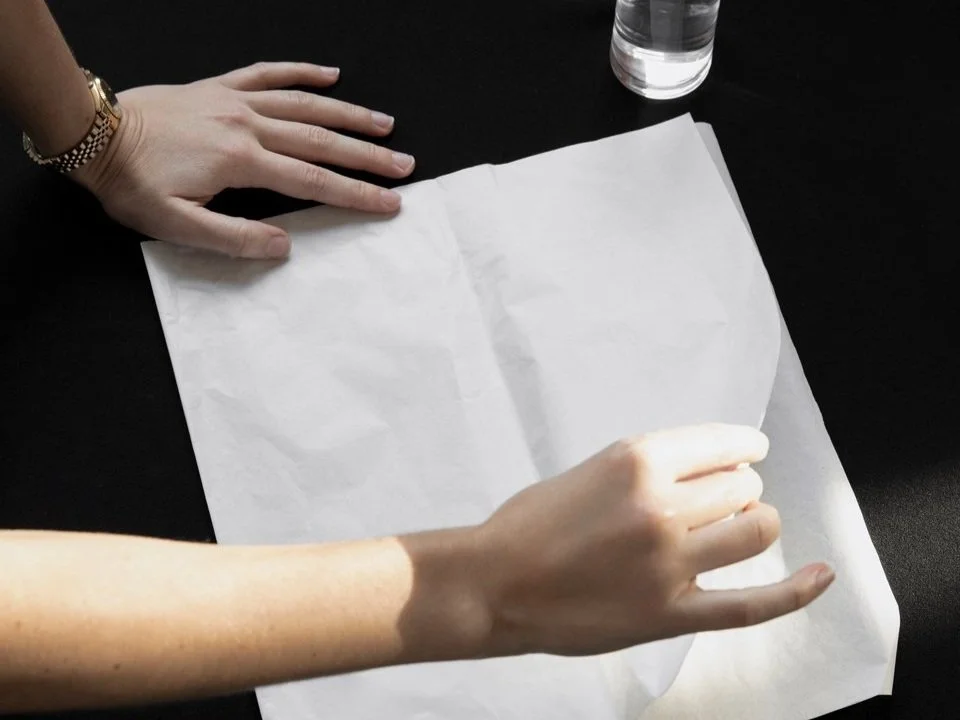 Two hands interact with a crumpled sheet of paper on a black surface, beside a glass of water.