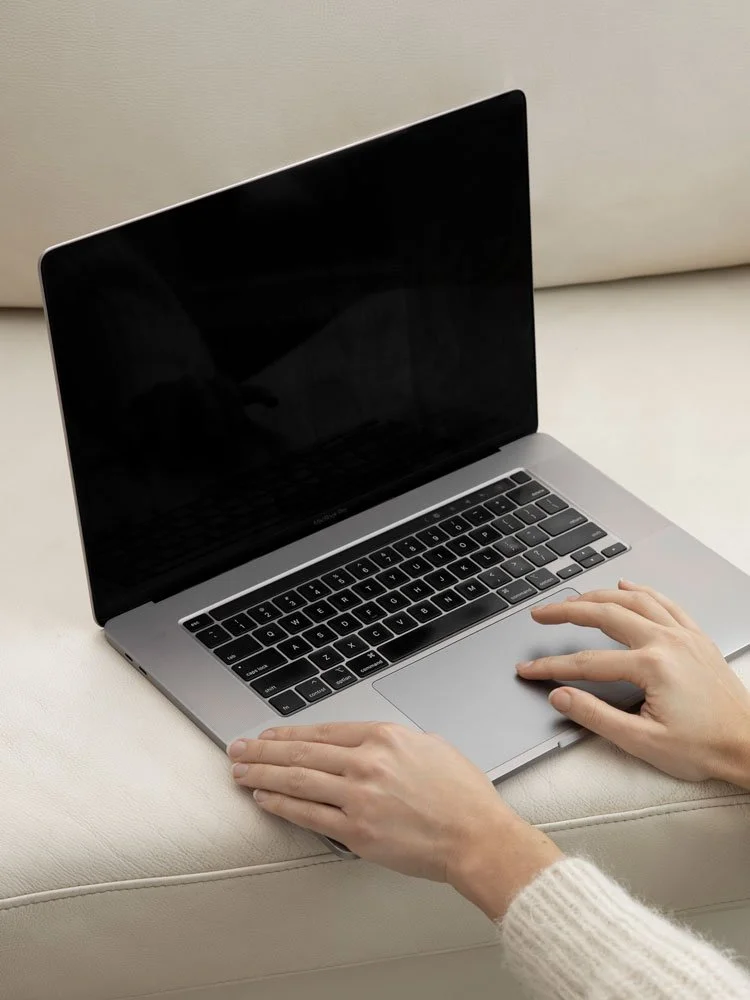 A person is using a silver laptop with a black keyboard on a white cushioned surface.