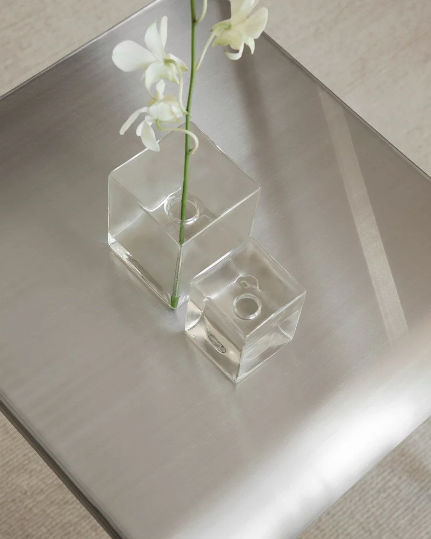 Minimal still life of two clear cube glass vases on a brushed metal table, one holding a white orchid stem.