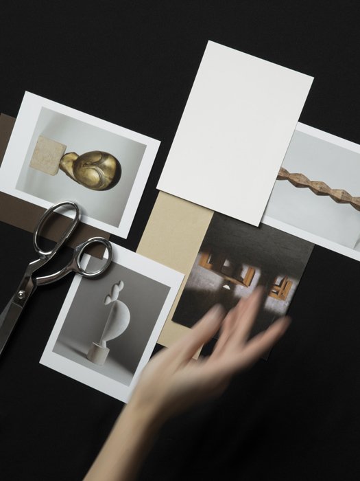 A flat lay of various photographs, some with sculptural objects, along with a pair of scissors and a person's hand reaching for one of the photos on a black surface.