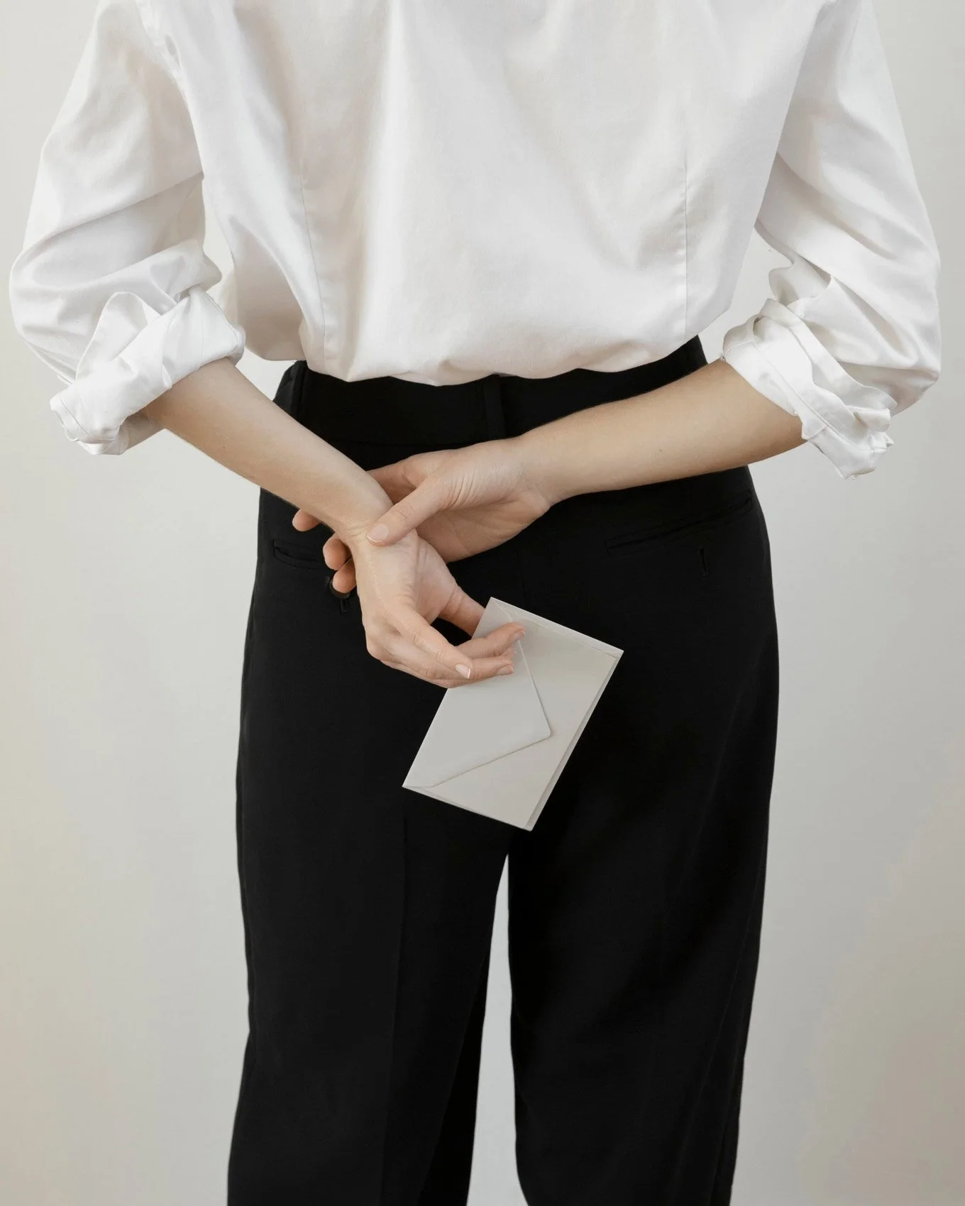 Person in a white shirt and black trousers holding a small white envelope behind their back.